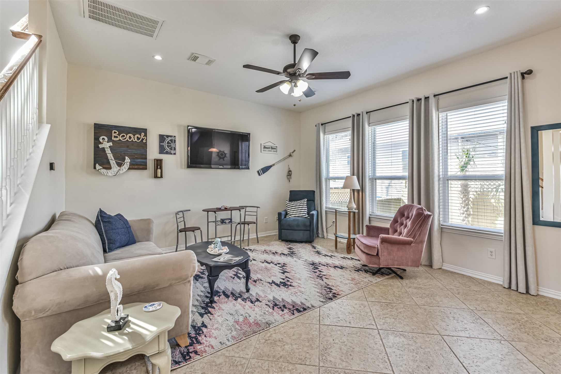 1815 28th Street Galveston, TX 77550 - Photo 26 of 42 This inviting living room features a cozy seating area with a mix of armchairs and a sofa, complemented by nautical-themed decor. The space is brightened by large windows with ample natural light and has a ceiling fan for comfort. The tiled floor and neutral walls create a welcoming atmosphere, perfect for relaxation or entertaining.