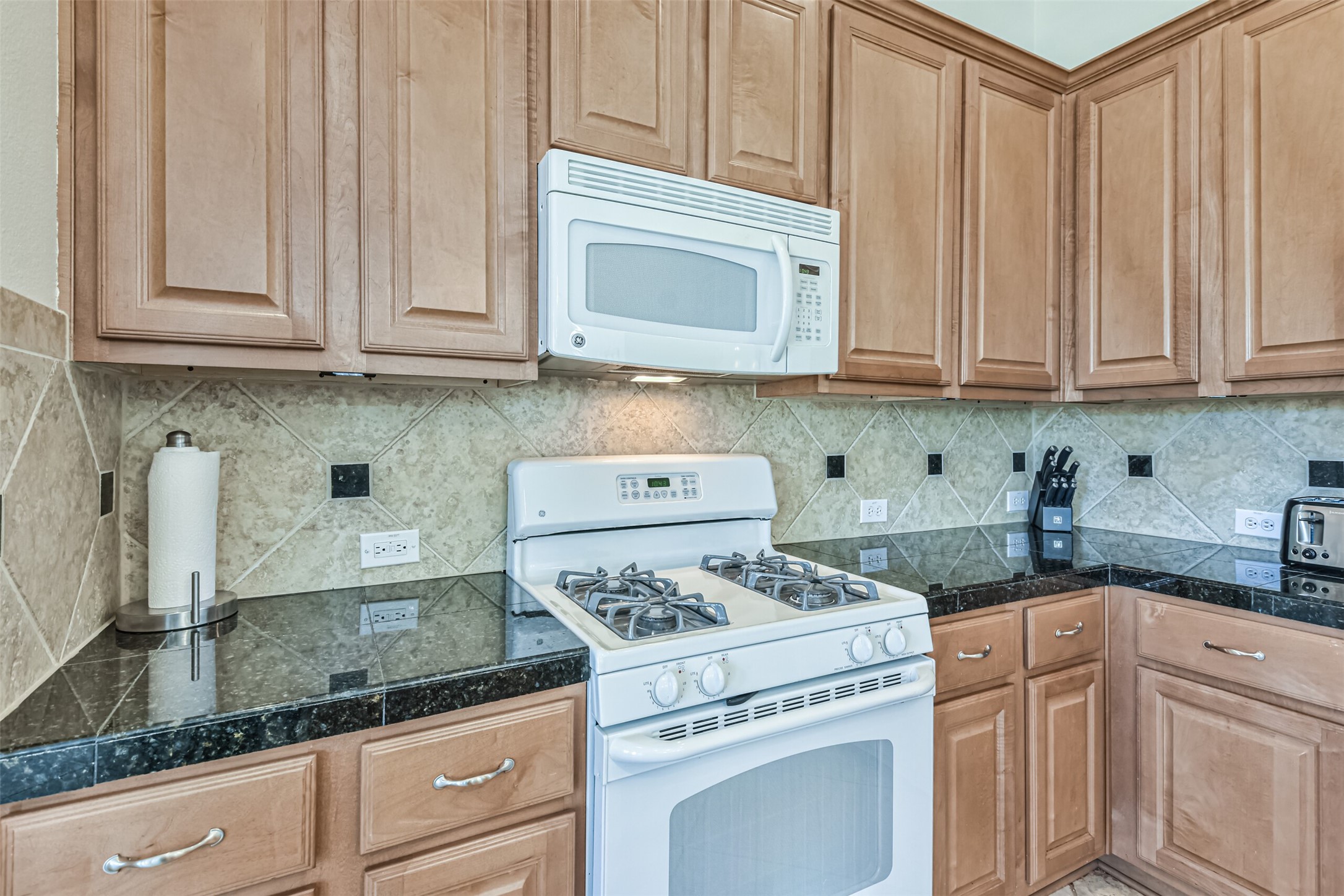 1815 28th Street Galveston, TX 77550 - Photo 28 of 42 This kitchen features wooden cabinetry, black granite countertops, and beige tile backsplash. It includes a white gas stove and microwave, with ample counter space and storage.