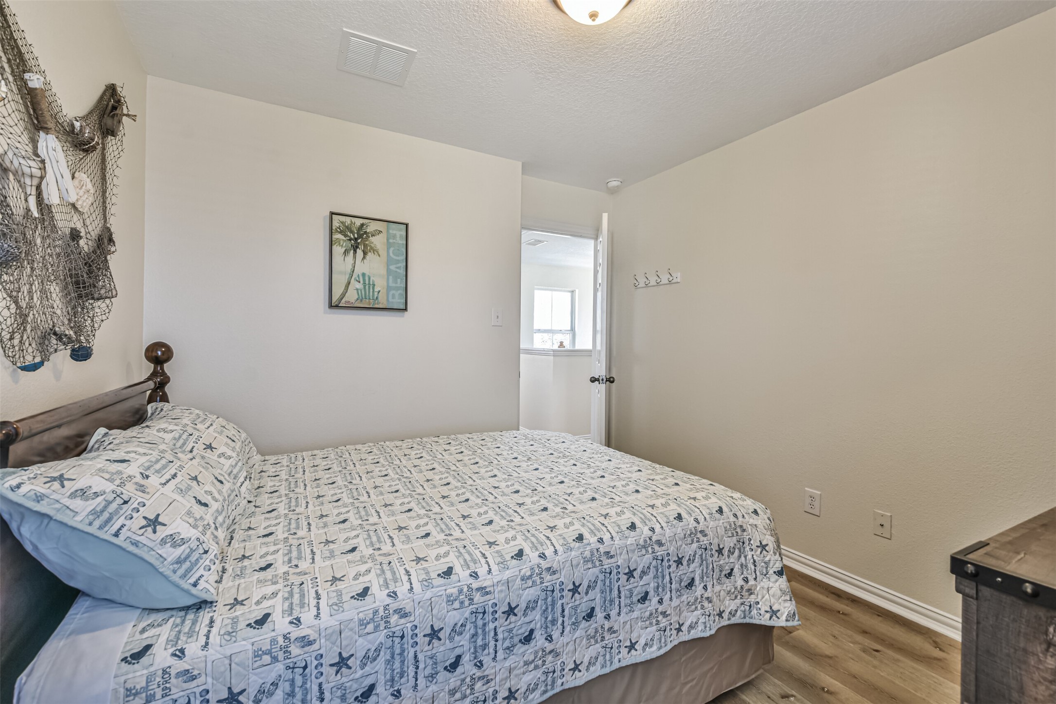 1815 28th Street Galveston, TX 77550 - Photo 35 of 42 Cozy bedroom with neutral walls and wood flooring, featuring a bed with a nautical-themed bedspread. Decor includes a beach-themed picture and wall-mounted hooks. Bright with natural light from an adjacent room.