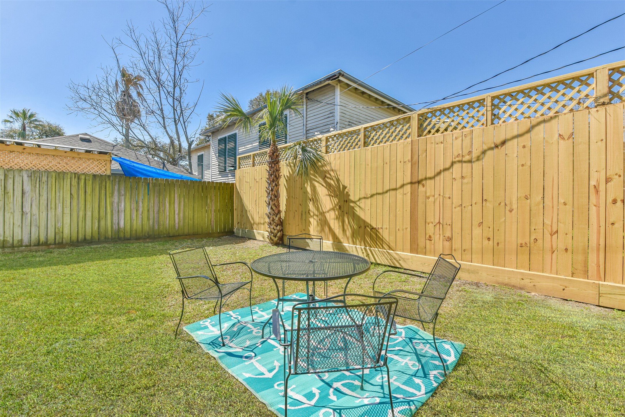 1815 28th Street Galveston, TX 77550 - Photo 36 of 42 This photo showcases a cozy backyard with a well-maintained lawn, a wooden privacy fence, and a small patio area featuring a round metal table and chairs on a decorative mat, perfect for outdoor gatherings.