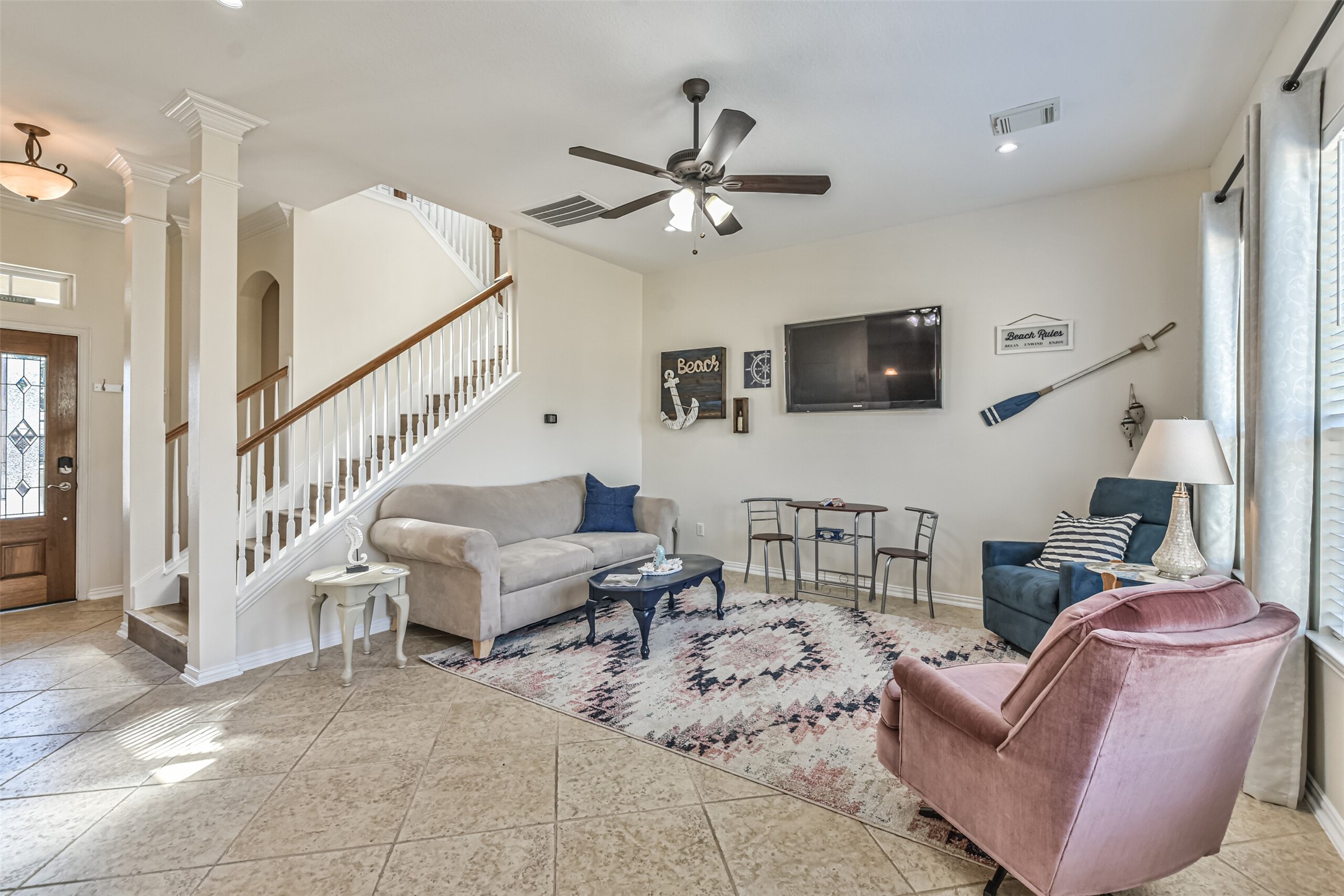 1815 28th Street Galveston, TX 77550 - Photo 4 of 42 This inviting living room features tile flooring, a cozy seating area with a couch and armchairs, and a mounted TV. A staircase with a wooden railing adds architectural interest. Natural light fills the space through large windows, enhancing the warm and welcoming atmosphere.