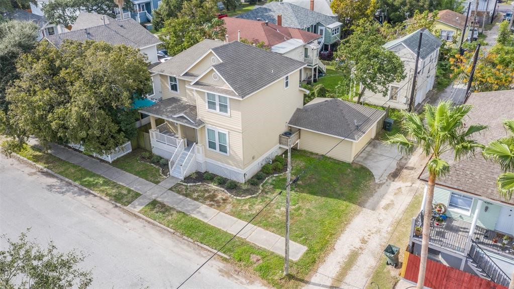 1815 28th Street Galveston, TX 77550 - Photo 41 of 42 This photo shows a two-story, light-colored house with a covered front porch and driveway. It's situated on a corner lot in a residential neighborhood, surrounded by trees and other homes. The property has a small front yard and a detached garage.