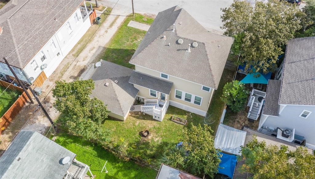 1815 28th Street Galveston, TX 77550 - Photo 42 of 42 This aerial photo shows a two-story home with a gabled roof and a detached garage. The backyard features a small lawn, trees, and a paved pathway leading to the back entrance. The surrounding area includes neighboring houses and a quiet street.