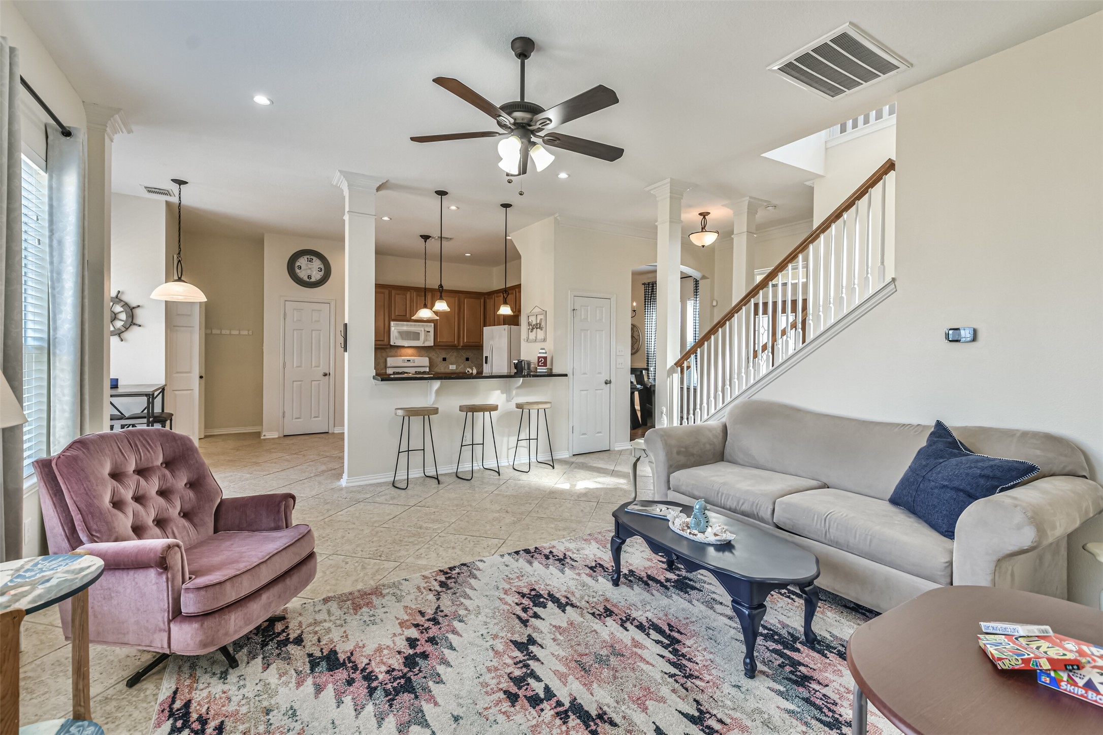 1815 28th Street Galveston, TX 77550 - Photo 5 of 42 This open-concept living area features a cozy seating arrangement with a plush armchair and sofa, a ceiling fan, and a stylish area rug. The space flows seamlessly into a modern kitchen with bar seating, pendant lighting, and wood cabinetry. A staircase leads to the upper level, enhancing the home's spacious feel.