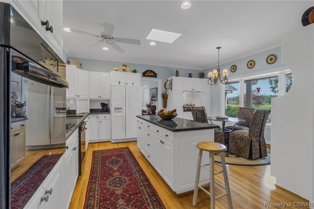 181 Duckbill Road White Stone, VA 22578 - Photo 23 of 50 Kitchen featuring black oven, a kitchen island, white fridge with ice dispenser, a skylight, dark countertops, white cabinets, ornamental molding, dishwashing machine, and light hardwood floors