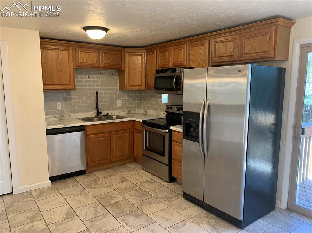 1057 Peterson Road Colorado Springs, CO 80915 - Photo 2 of 32 Kitchen with stainless steel appliances, decorative backsplash, brown cabinetry, and a textured ceiling