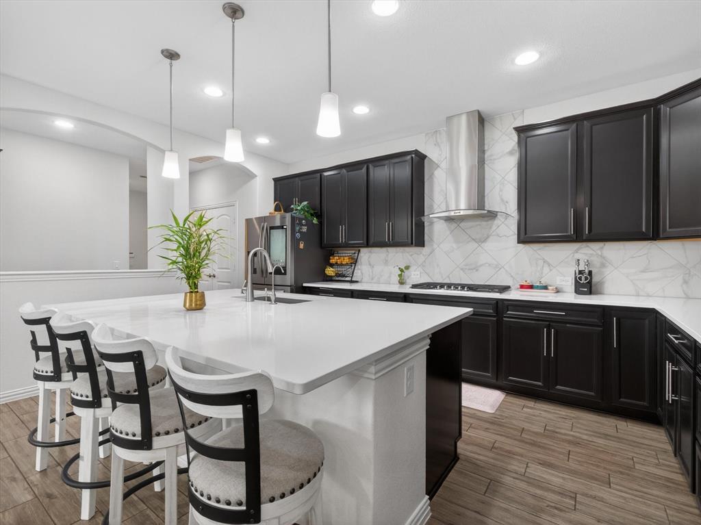636 Corner Post Path Celina, TX 75009 - Photo 10 of 40 Contemporary kitchen featuring dark cabinetry, a white countertop island with integrated sink, stainless steel appliances, a range hood, and wood-finish flooring