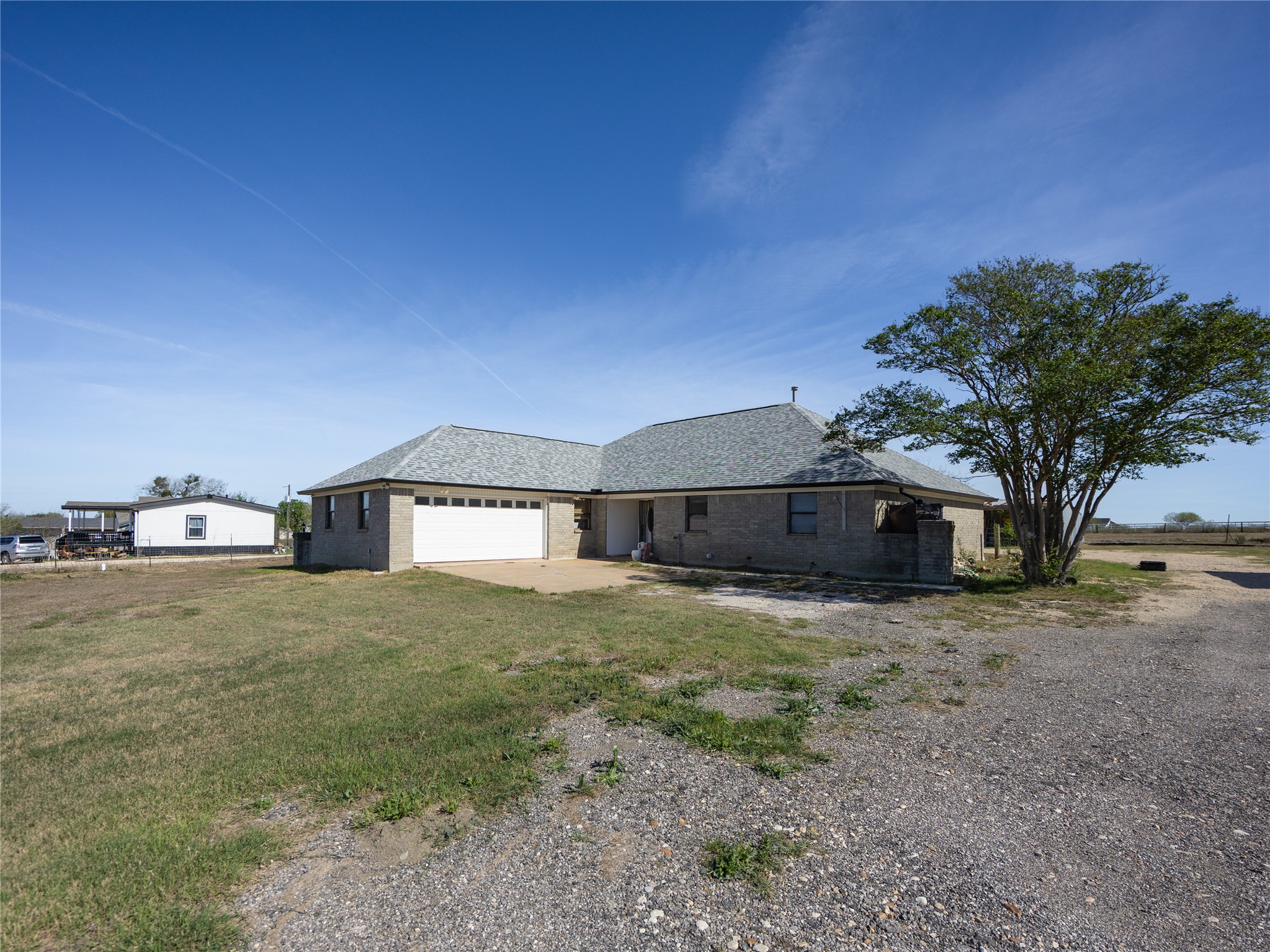 2412 Williamson Road Lockhart, TX 78644 - Photo 14 of 36 Single-story residence featuring a gray brick exterior, a light gray shingle roof, an attached two-car garage with a white door, and a mature tree providing shade