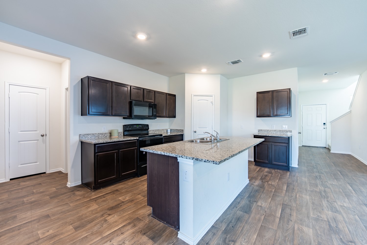 14917 Bajada Rd Manor Manor, TX 78653 - Photo 12 of 31 Open-concept kitchen featuring dark wood cabinetry, light granite countertops, a central island with a double sink, black appliances, and wood-finish flooring