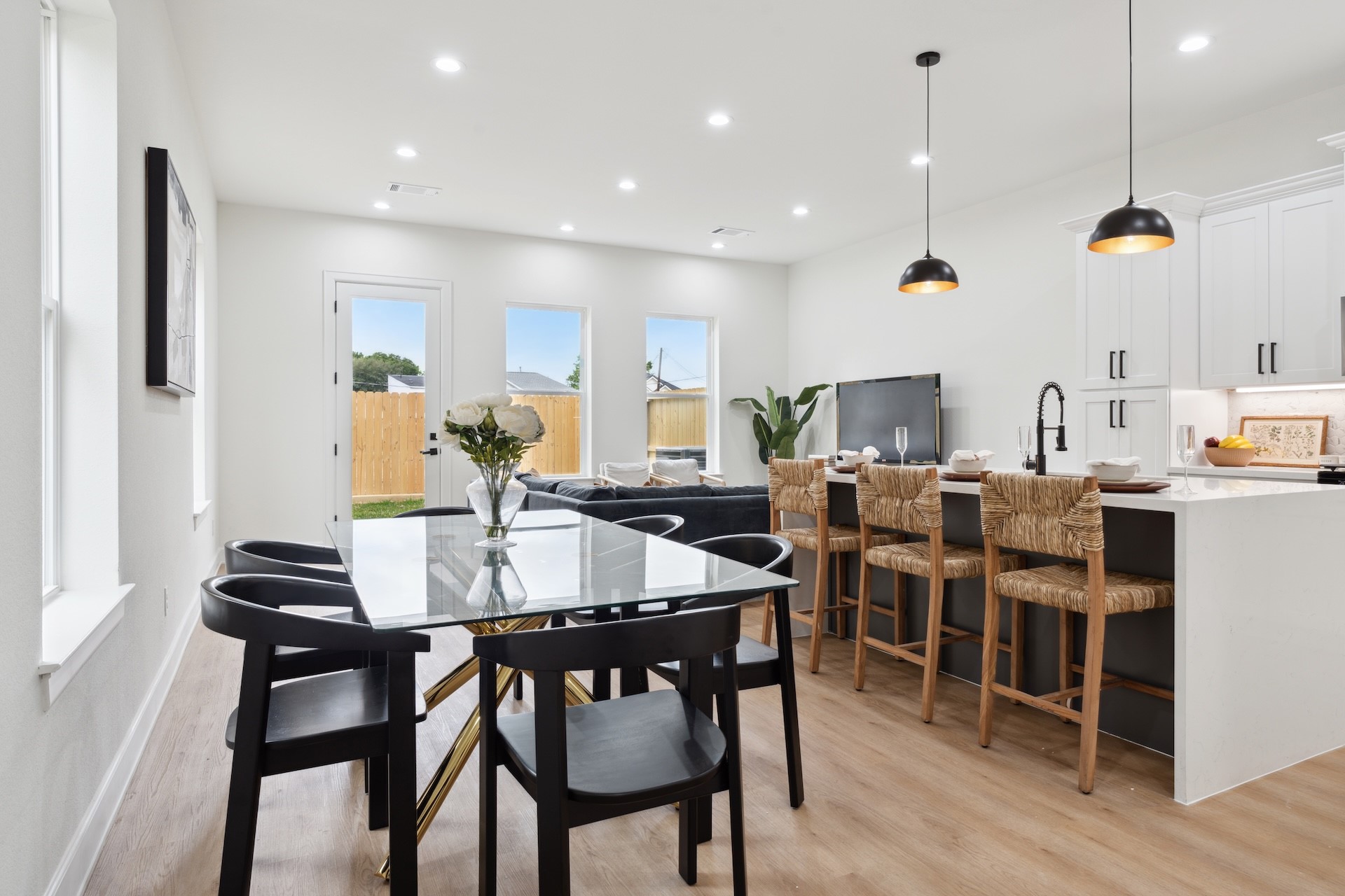 Light-filled open layout — formal dining area with glass table seating, island bar setup with woven counter stools, and direct back patio door access.