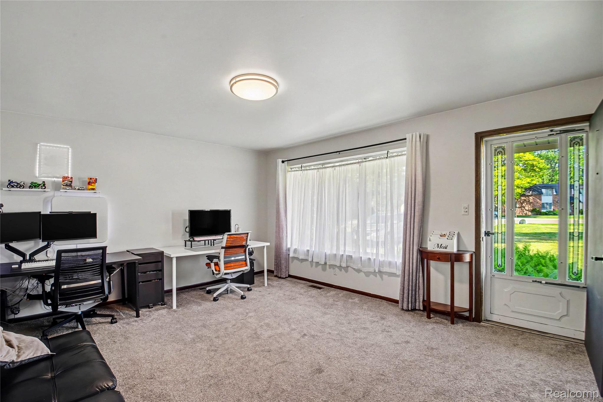 Spacious room featuring neutral carpeting, a large window with sheer and solid drapes, and a decorative front door with clear and green glass panels