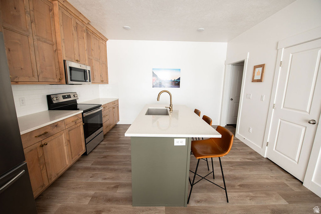 384 Ropelato Drive Nibley, UT 84321 - Photo 6 of 25 Kitchen featuring wood-finish cabinetry, stainless steel appliances, a subway tile backsplash, light-toned countertops, and a center island with a sink and brass-finish faucet