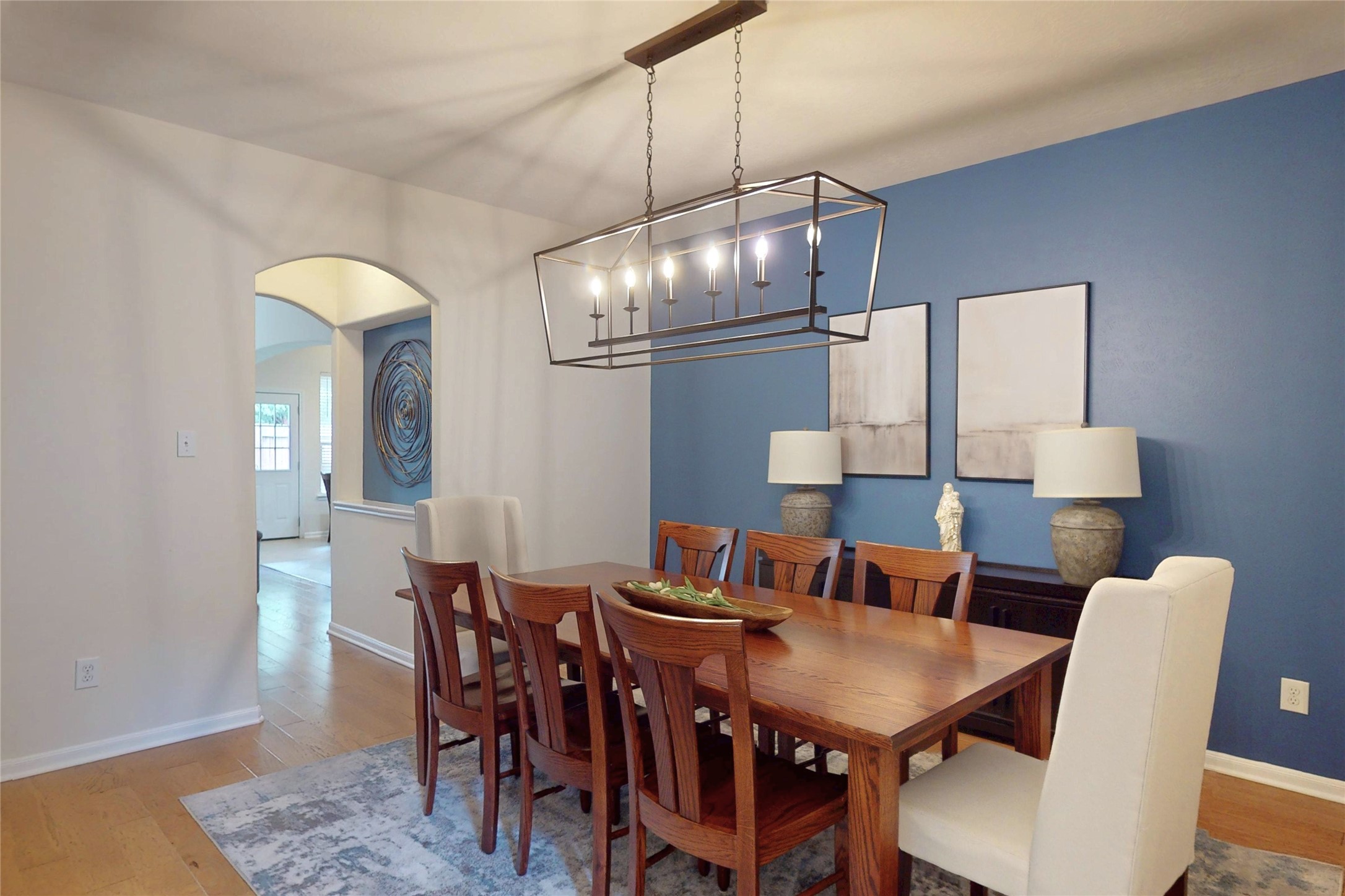 17402 Rainer Valley Lane Humble, TX 77346 - Photo 3 of 33 The photo showcases a modern dining room with a wooden table and chairs, set against a blue accent wall. It features contemporary lighting, abstract art, and a soft area rug, creating a welcoming and stylish atmosphere.