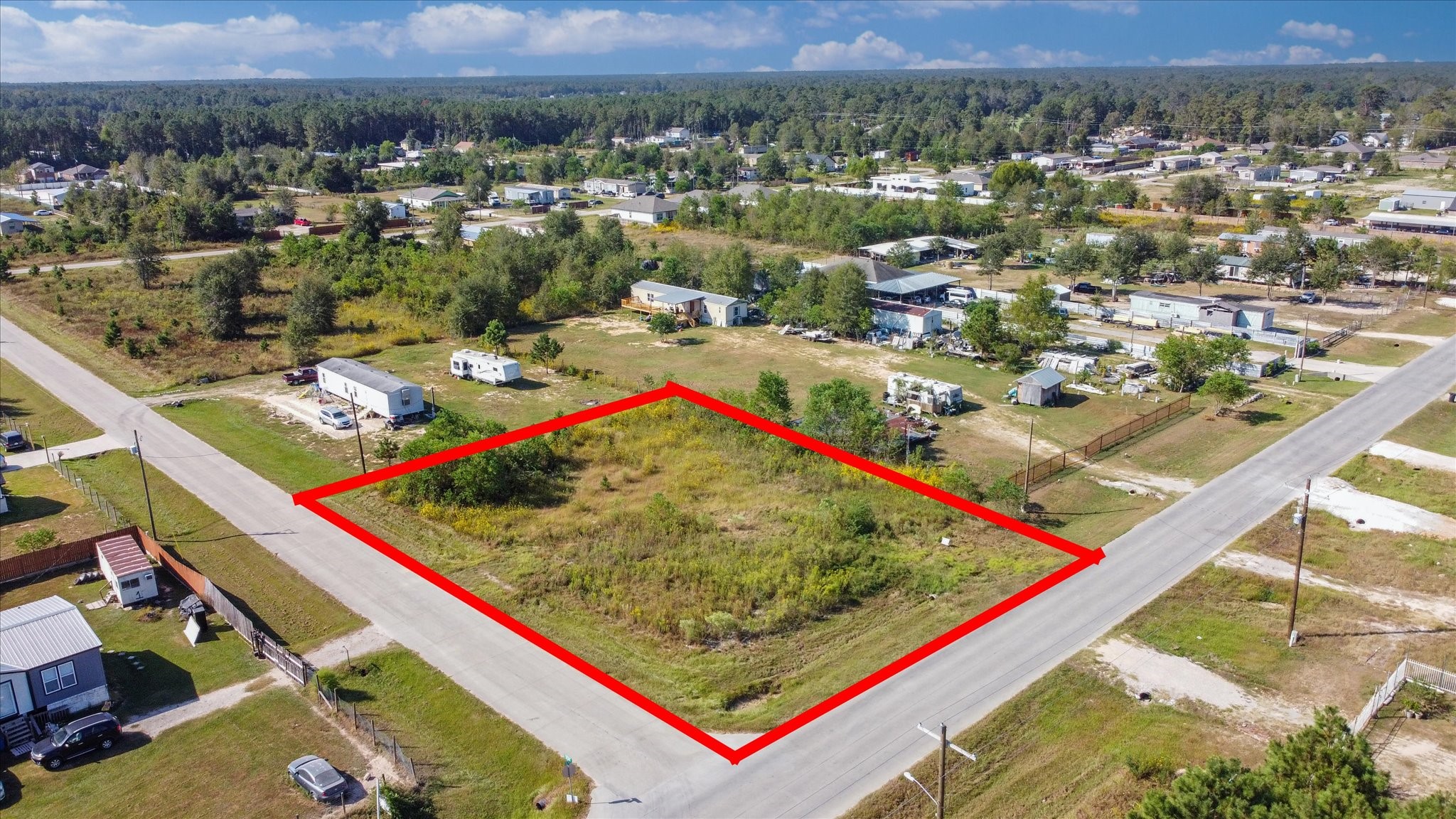 This is an aerial view of a vacant parcel of land outlined in red, located at a corner lot within a residential area with a mix of houses and mobile homes.