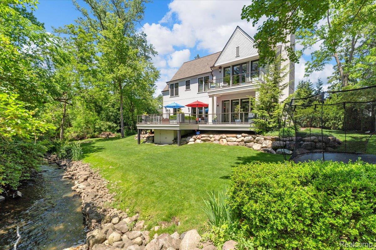 195 Baldwin Road Birmingham, MI 48009 - Photo 1 of 76 Rear exterior featuring a well-maintained lawn, multi-level deck with glass railings, an upper-level balcony, a natural stream, and extensive landscaping with mature trees