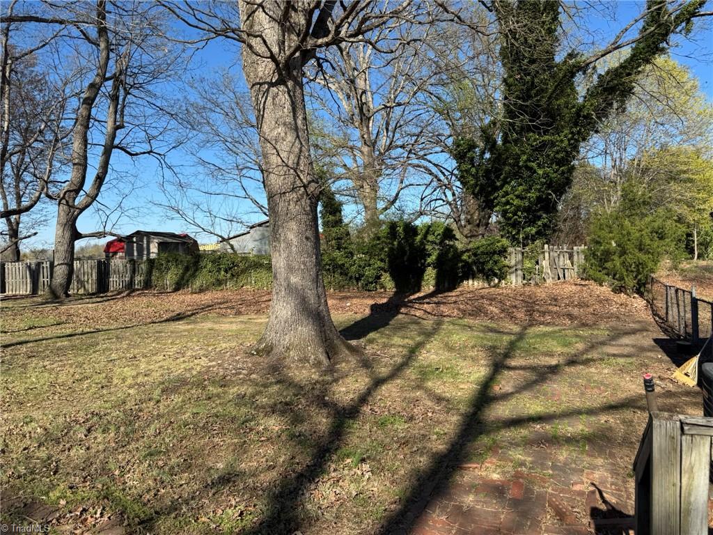 2023 Benaja Road Reidsville, NC 27320 - Photo 33 of 36 Side Yard with Fenced Area and Mature Trees at 2023 Benaja Road, Reidsville, NC 27320, Rockingham County. Side yard view showing fenced backyard area with mature hardwood trees. Wooden privacy fence and hedgerow along the property line. Open grassy space.