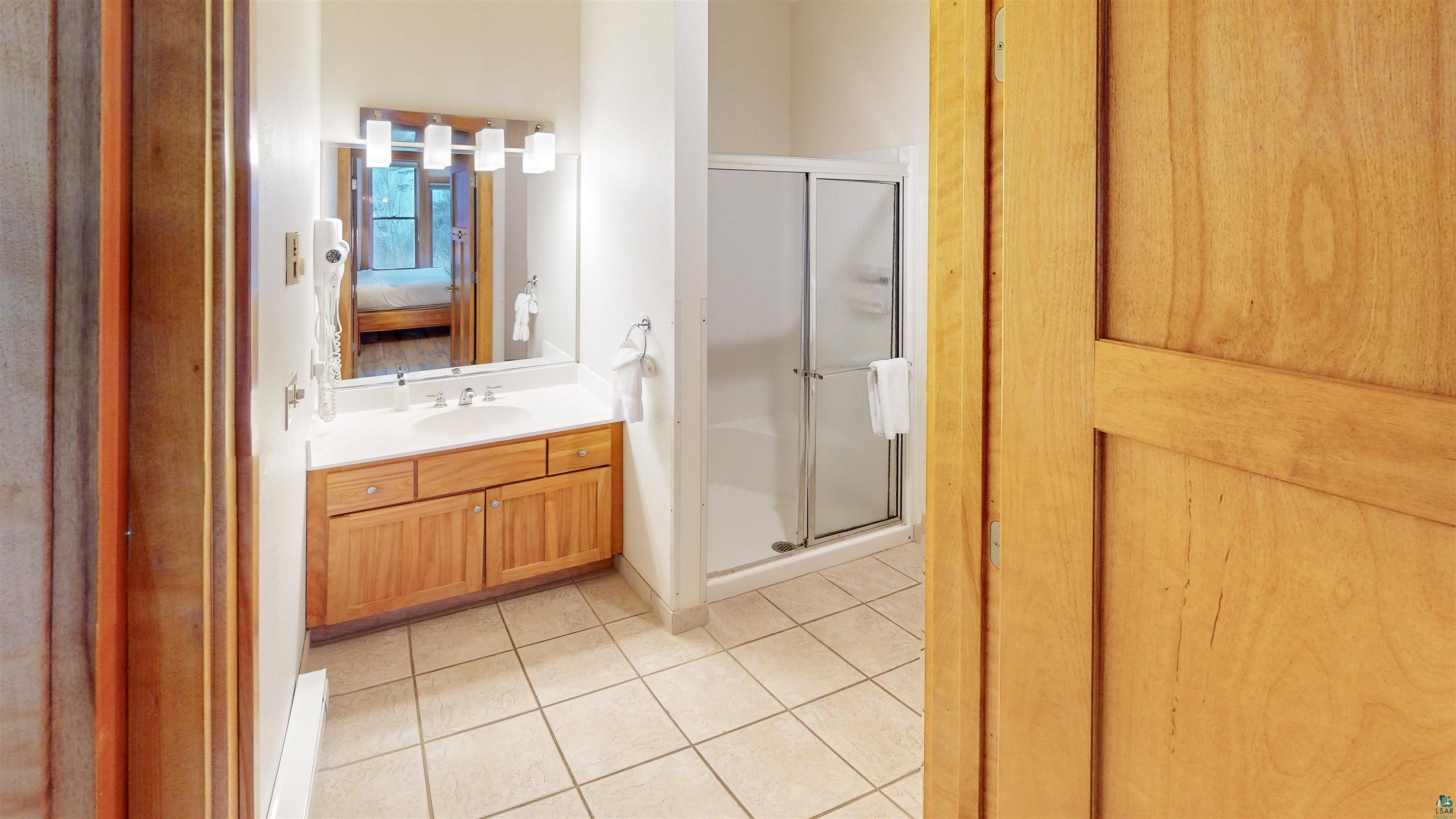 504-506 Poplar River Road, Unit 505 Lutsen, MN 55612 - Photo 24 of 39 Bathroom featuring a wood-finish vanity with an integrated sink, a large mirror with overhead lighting, a shower enclosure with sliding glass doors, and tiled flooring