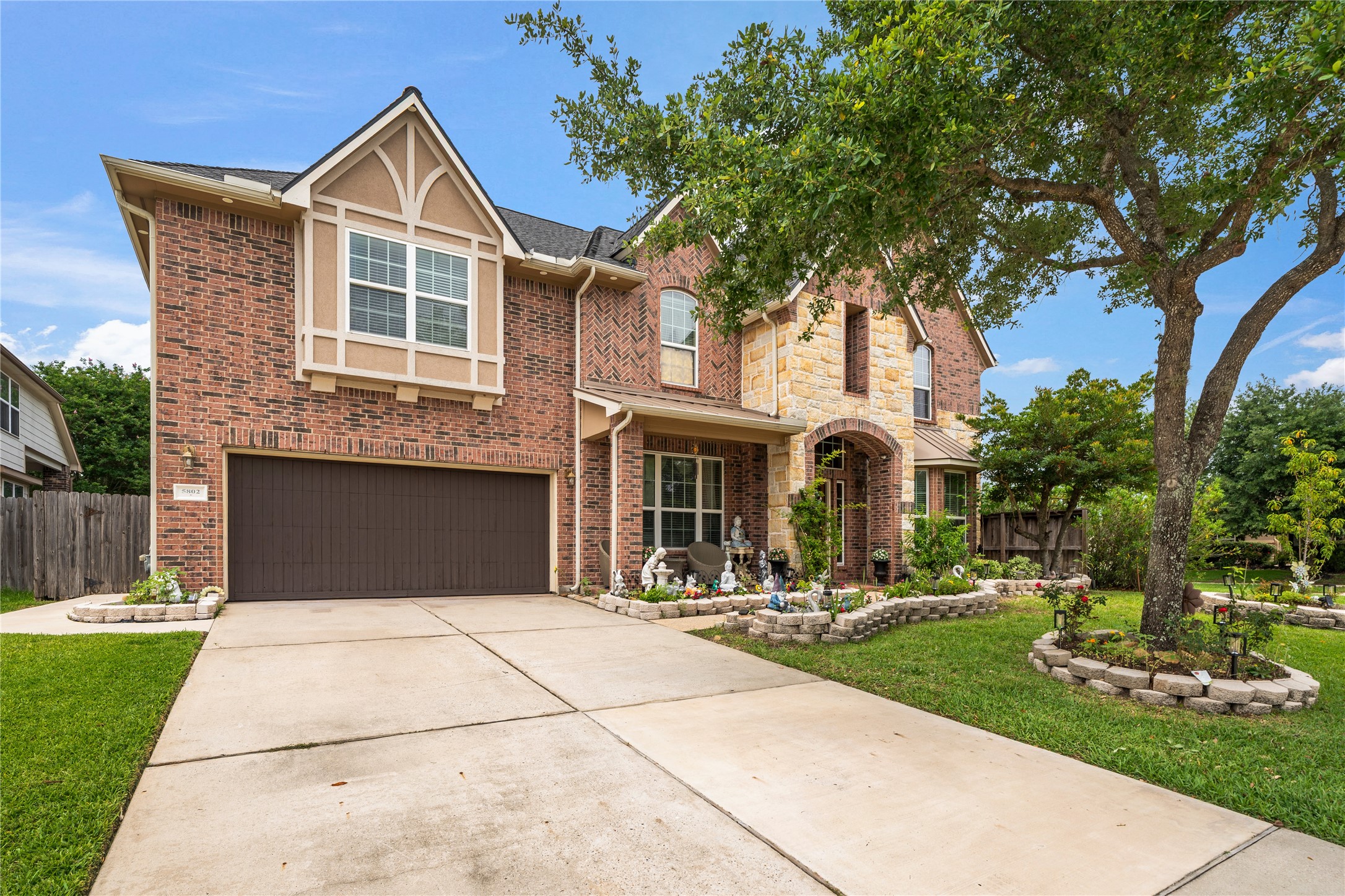 5802 Layton Meadows Lane Spring, TX 77379 - Photo 2 of 42 Front exterior with garage. Brick and stone two-story facade, dark wood garage door, stone-edged garden beds, manicured lawn, mature oak tree.