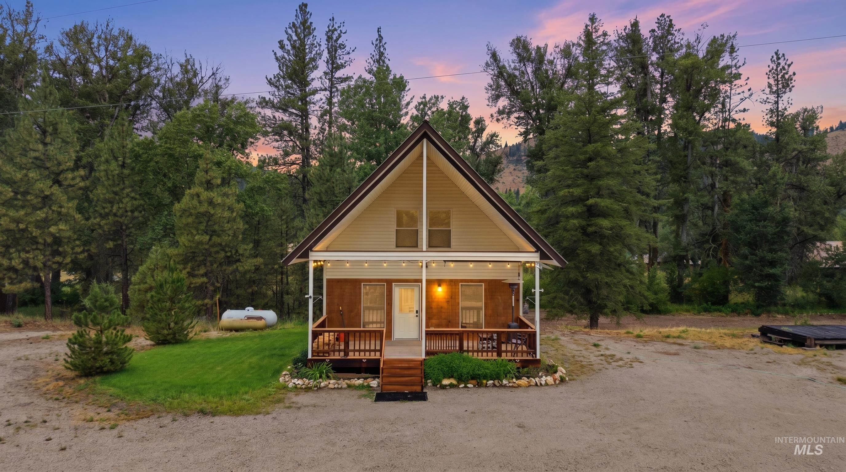 3980 North Pine-Featherville Road Mountain Home, ID 83647 - Photo 1 of 35 A-frame cabin featuring a covered front porch with string lighting, a gravel driveway, and a tree-lined backdrop
