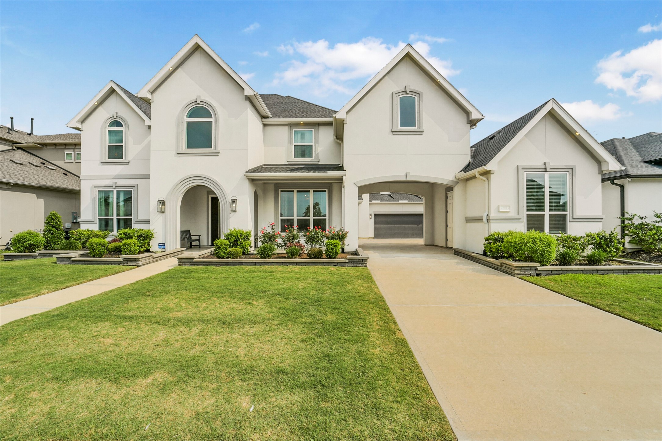 Modern, two-story house with a clean white facade and arched entryway. The home includes a well-maintained front lawn, manicured shrubs, and a spacious driveway leading to a covered car port. The architecture is elegant, with large windows providing ample natural light.