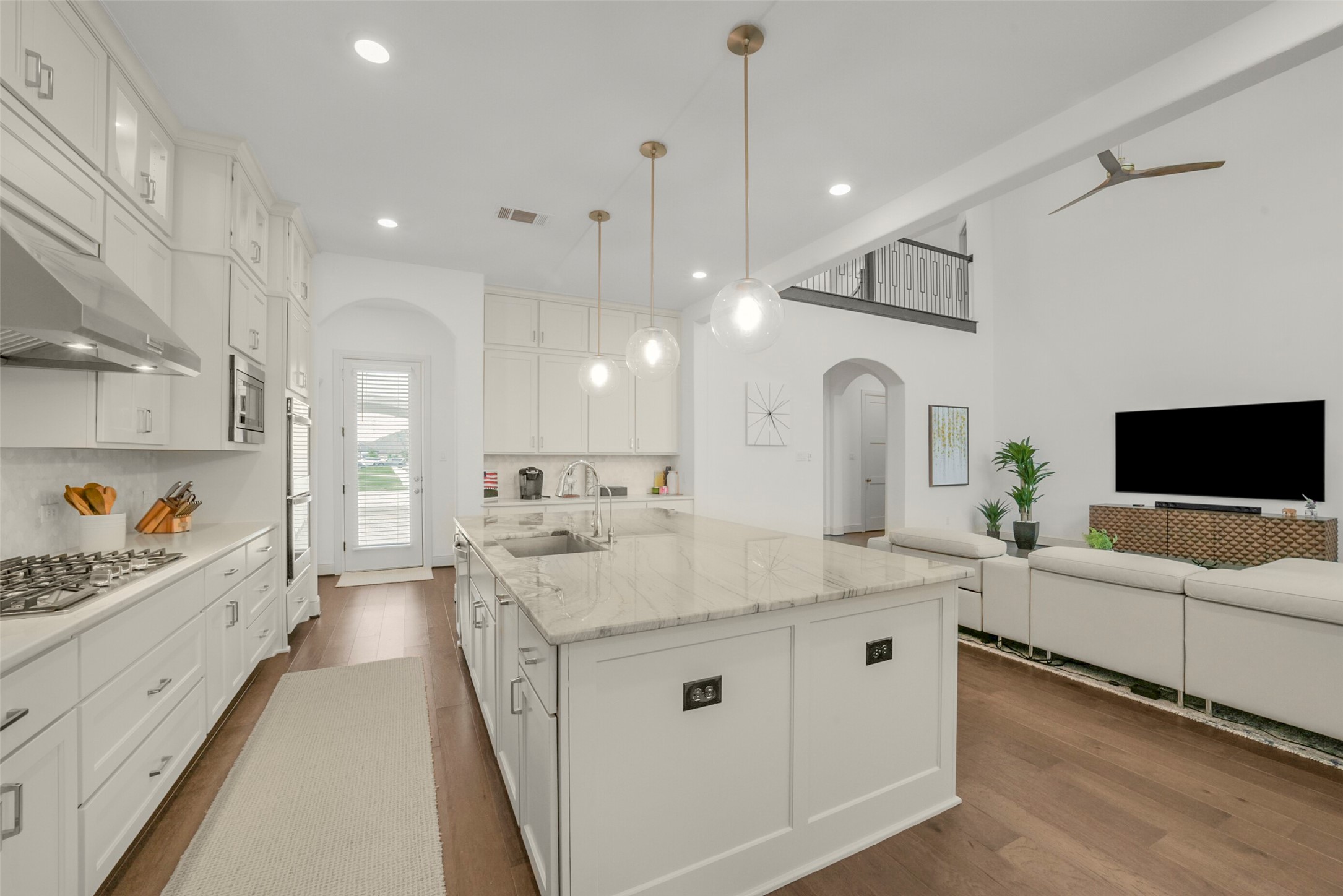2018 Grove Terrace Lane Manvel, TX 77578 - Photo 10 of 50 Modern, open-concept kitchen and living area with high ceilings. The kitchen features white cabinetry, a large marble island, and stainless steel appliances. The space is bright with recessed lighting and pendant lights, and it opens into a cozy living area with a sectional sofa and a wall-mounted TV. The hardwood floors add warmth to the sleek design.