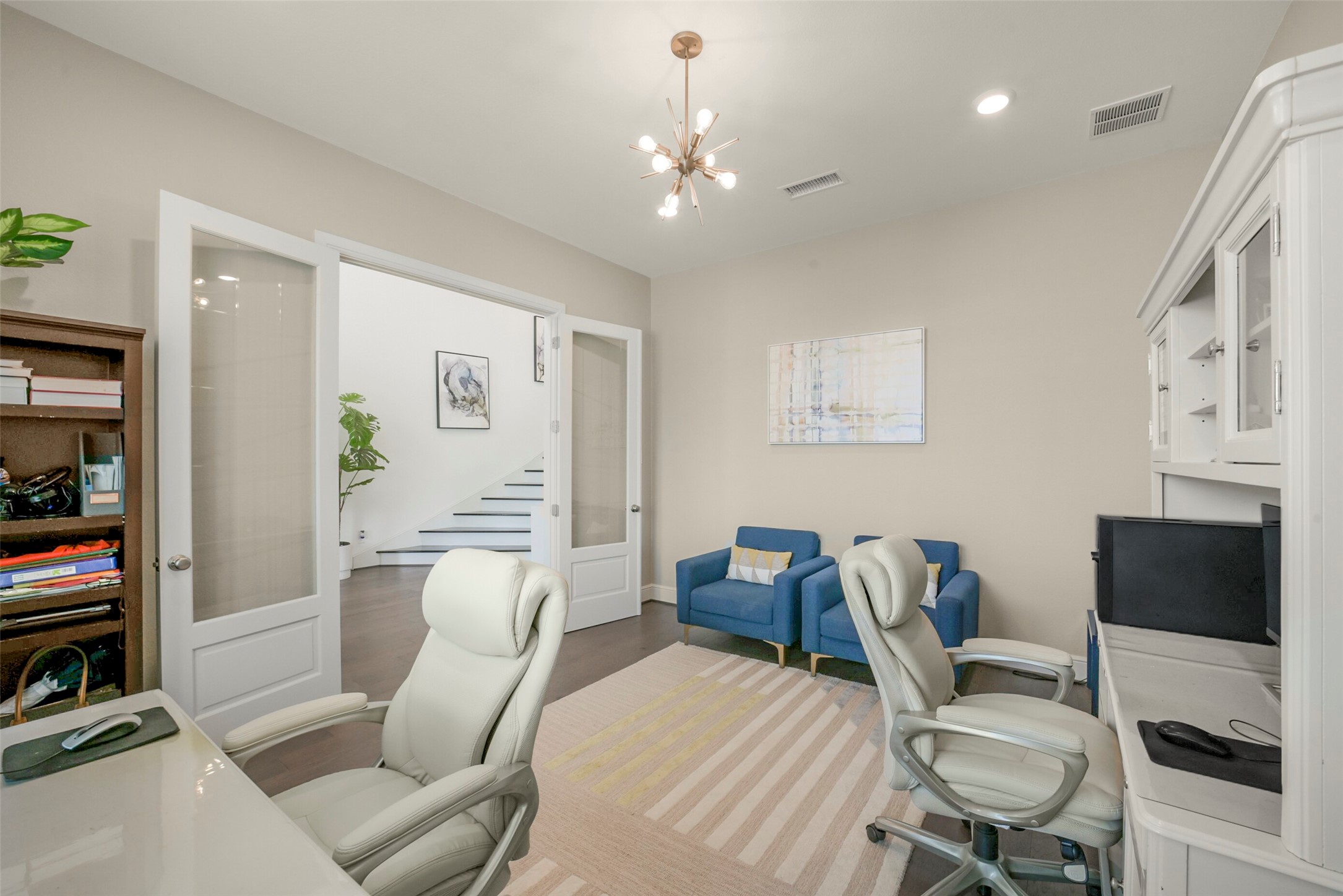 2018 Grove Terrace Lane Manvel, TX 77578 - Photo 20 of 50 Bright home office with modern furnishings, including a desk with two chairs, a computer setup, and built-in shelving. The room features a stylish light fixture, glass-paneled doors, and a cozy seating area with two blue chairs. There's a view of the staircase and entryway, enhancing the open feel.