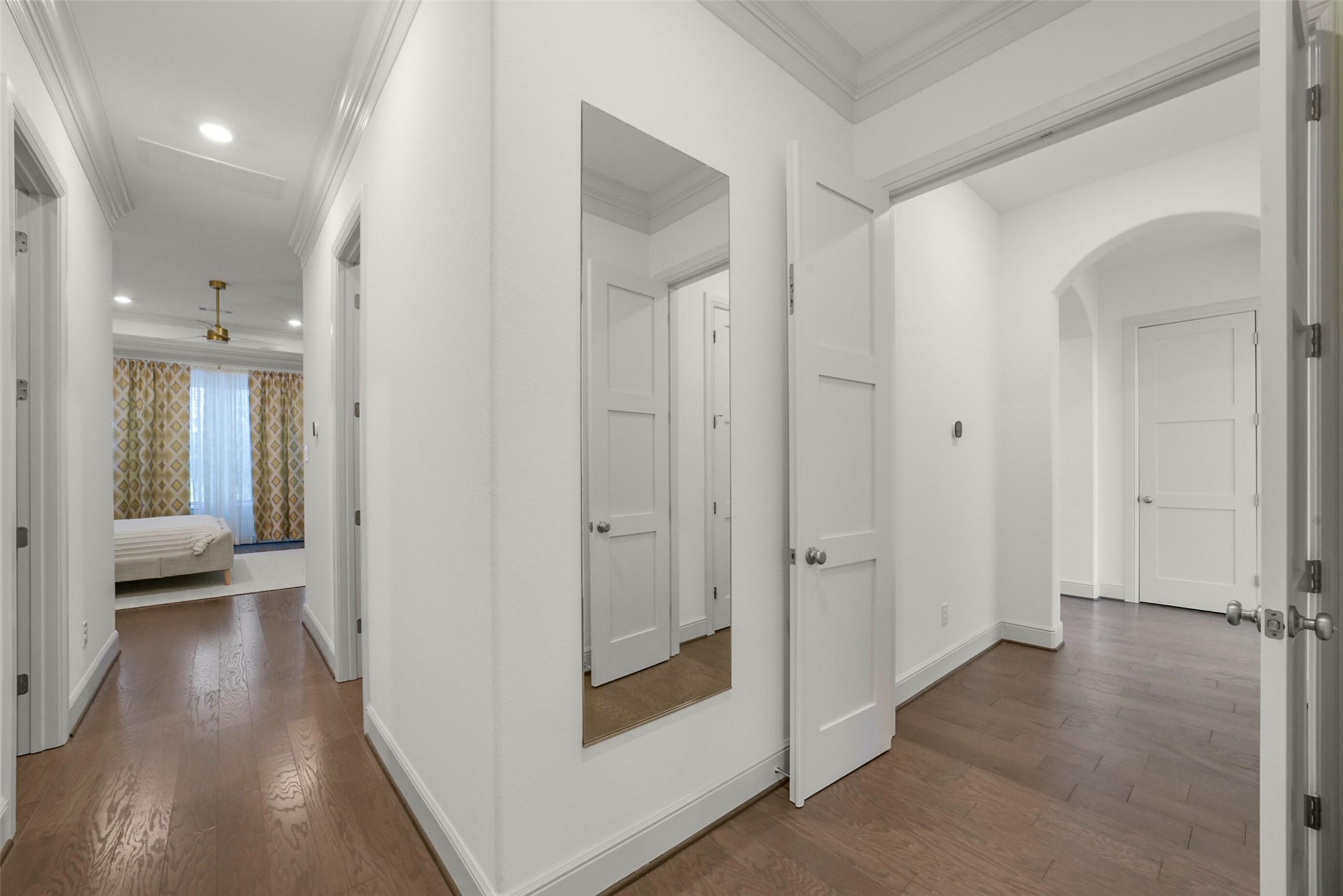 2018 Grove Terrace Lane Manvel, TX 77578 - Photo 22 of 50 This photo shows a bright, modern hallway leading to the primary bedroom with polished hardwood floors and white walls. There are multiple doors leading to the primary bedroom and primary spa like bathroom, including a glimpse of large windows and patterned curtains. A wall-mounted mirror adds depth to the space.