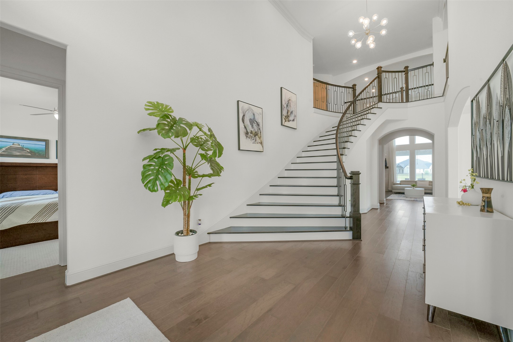 2018 Grove Terrace Lane Manvel, TX 77578 - Photo 3 of 50 Bright, spacious entryway featuring a sweeping staircase with elegant iron railings, hardwood floors, and modern decor. There's a glimpse of a cozy bedroom on the left and a well-lit living area with large windows straight ahead.
