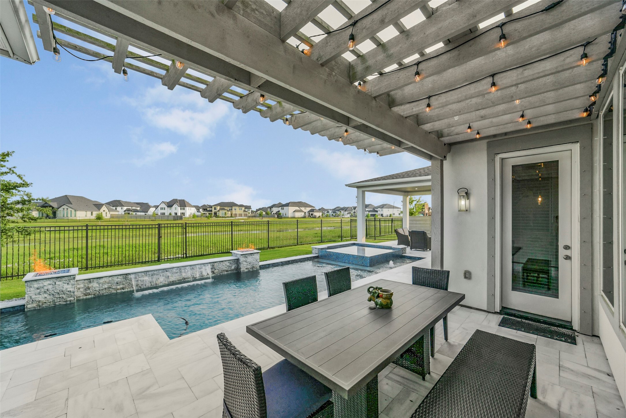 2018 Grove Terrace Lane Manvel, TX 77578 - Photo 41 of 50 Modern outdoor patio with a pergola and string lights, overlooking a sleek pool and spa. The space includes a dining area with a table and chairs, perfect for entertaining, and offers scenic views of a spacious, open green area beyond the fence.