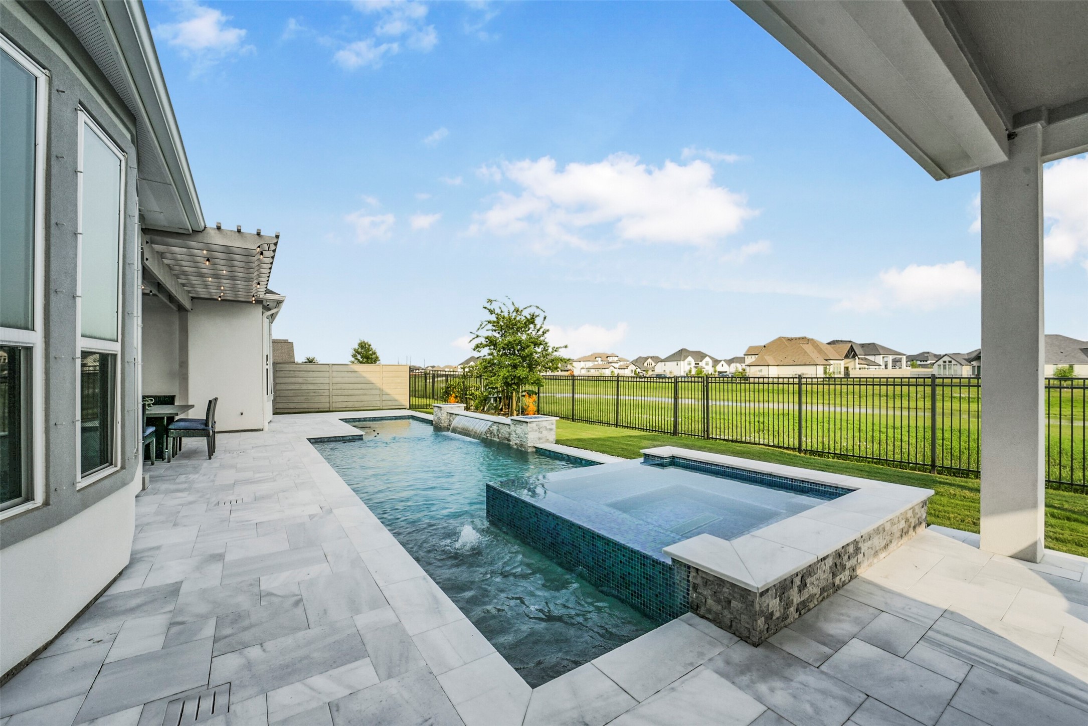2018 Grove Terrace Lane Manvel, TX 77578 - Photo 43 of 50 Modern backyard featuring a sleek pool with a spa, surrounded by a spacious, tiled patio. The area is enclosed by an iron fence, offering views of a well-maintained lawn, creating a serene and inviting outdoor space.