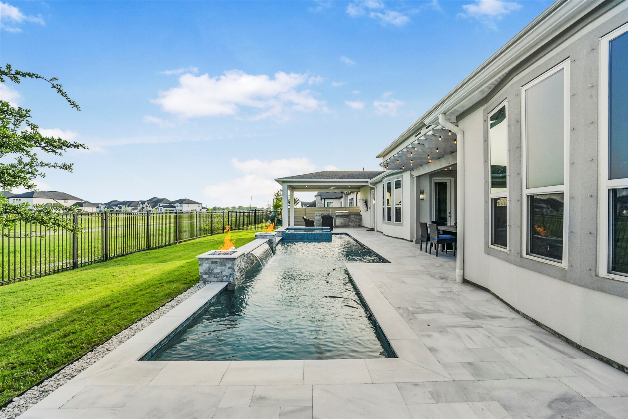 2018 Grove Terrace Lane Manvel, TX 77578 - Photo 44 of 50 Modern home with a sleek backyard featuring a long, narrow pool and fire features. The patio area is spacious with elegant stone flooring, and there's a covered outdoor dining space. The yard is fenced, offering a view of a lush green field beyond.