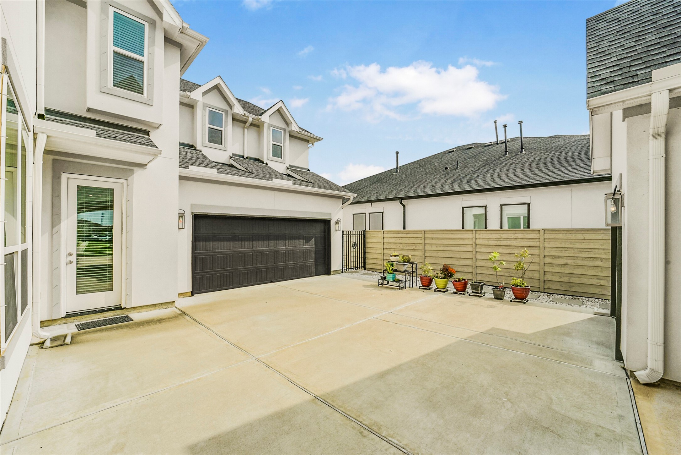 2018 Grove Terrace Lane Manvel, TX 77578 - Photo 49 of 50 Spacious, paved courtyard with a modern white exterior and a two-car garage. There's a wooden fence lined with potted plants, adding a touch of greenery. The area is well-lit and appears ideal for outdoor activities or parking.