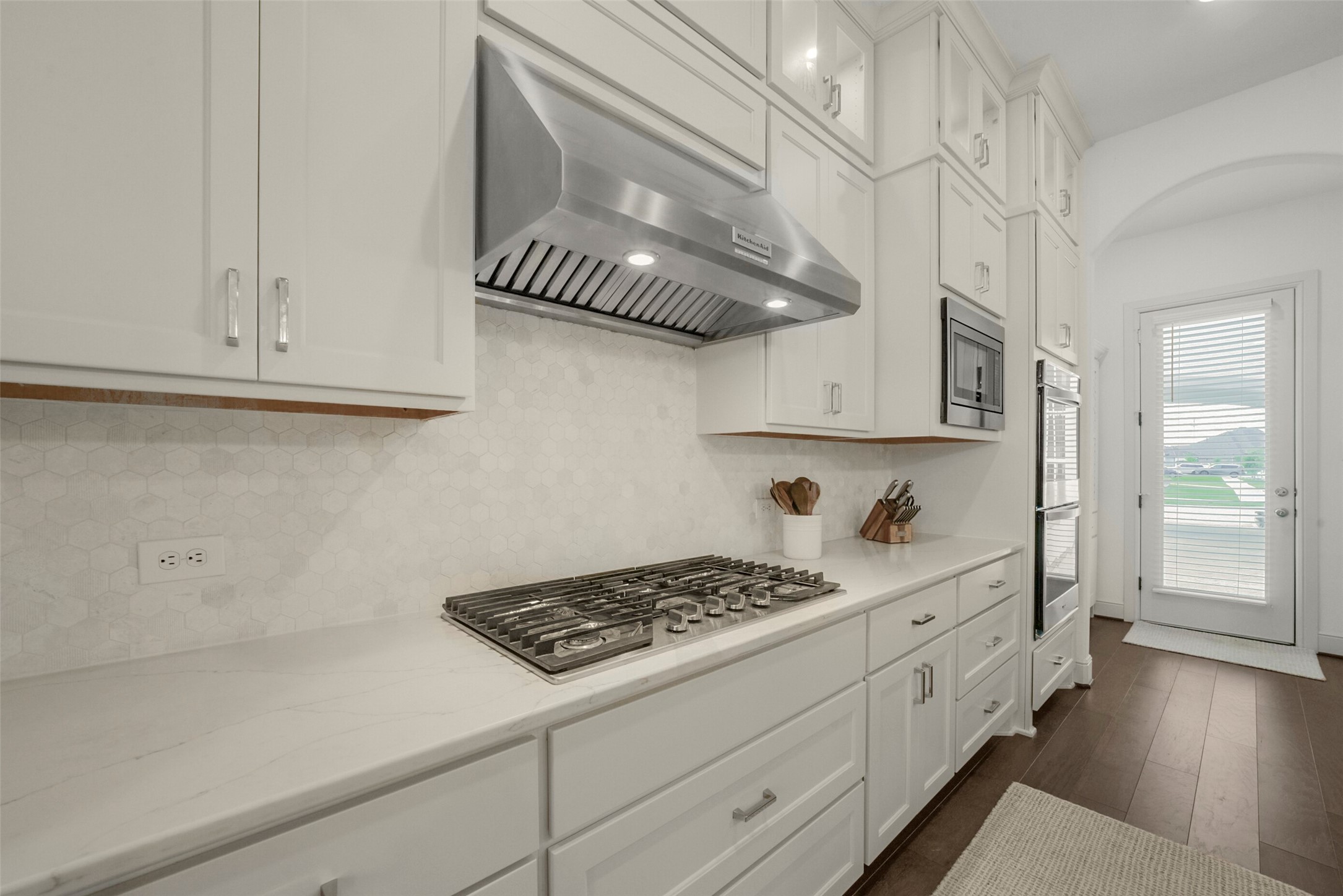 2018 Grove Terrace Lane Manvel, TX 77578 - Photo 8 of 50 This kitchen features sleek, white cabinetry with modern hardware, a built-in gas cooktop with a stainless steel hood, and a clean, hexagonal tile backsplash. It includes integrated appliances and has a bright, airy feel with ample natural light from the nearby door.