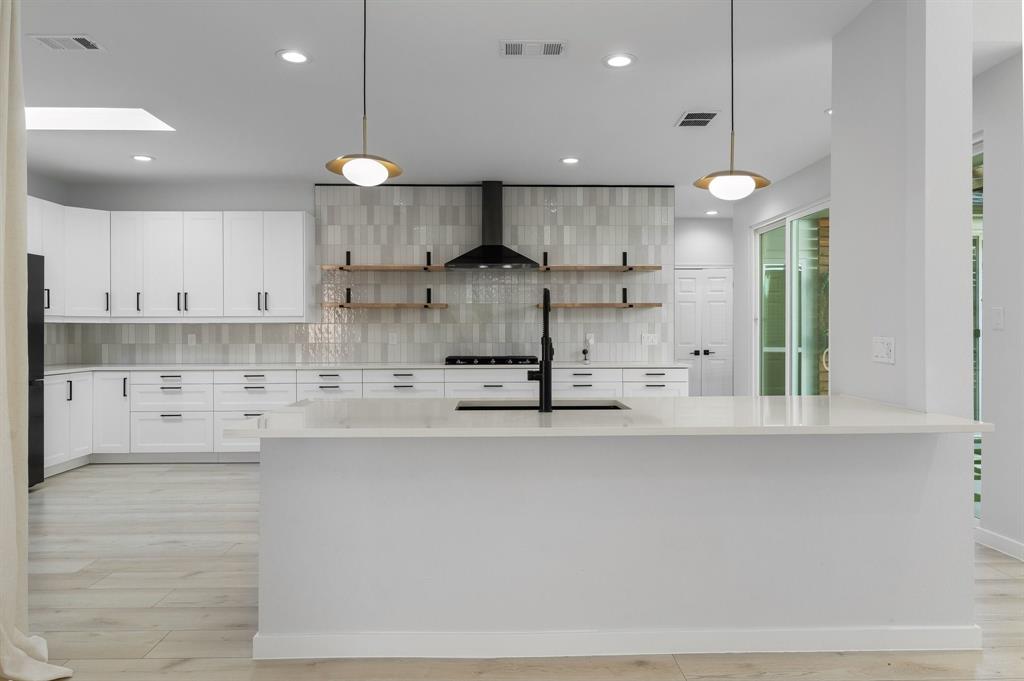 14140 Regency Place Dallas, TX 75254 - Photo 15 of 38 Modern kitchen featuring a large central island with a built-in sink and matte black faucet, extensive white cabinetry with black hardware, a light-toned tile backsplash, and wood-finish open shelving
