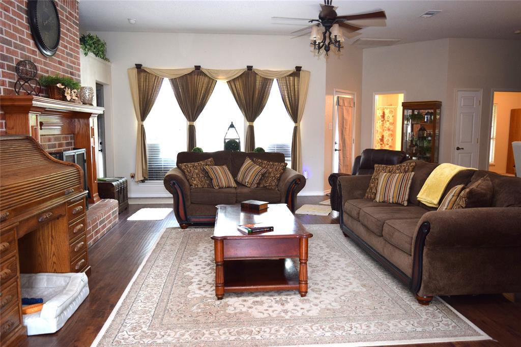 4201 Glistening Spring Rowlett, TX 75088 - Photo 5 of 33 Spacious living area featuring wood-finish flooring, a brick fireplace with a wood mantel, multiple windows with blinds, a ceiling fan with integrated lighting, and neutral wall tones