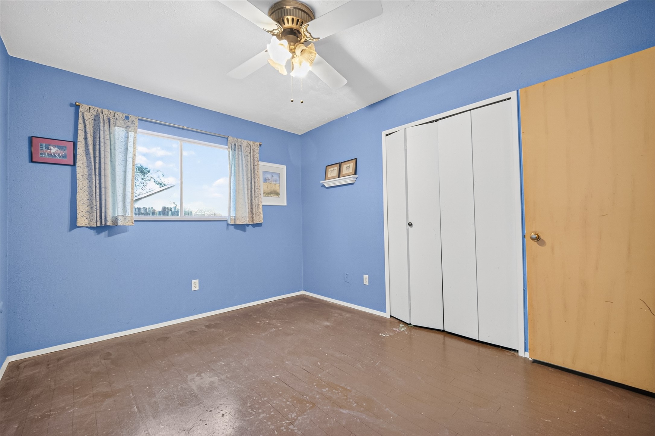 22926 Millgate Drive Spring, TX 77373 - Photo 21 of 34 This room features blue walls, a ceiling fan with a light, and a window with curtains that brightens the space. It has a closet with white folding doors and a simple brown floor, making it a versatile space for various uses.