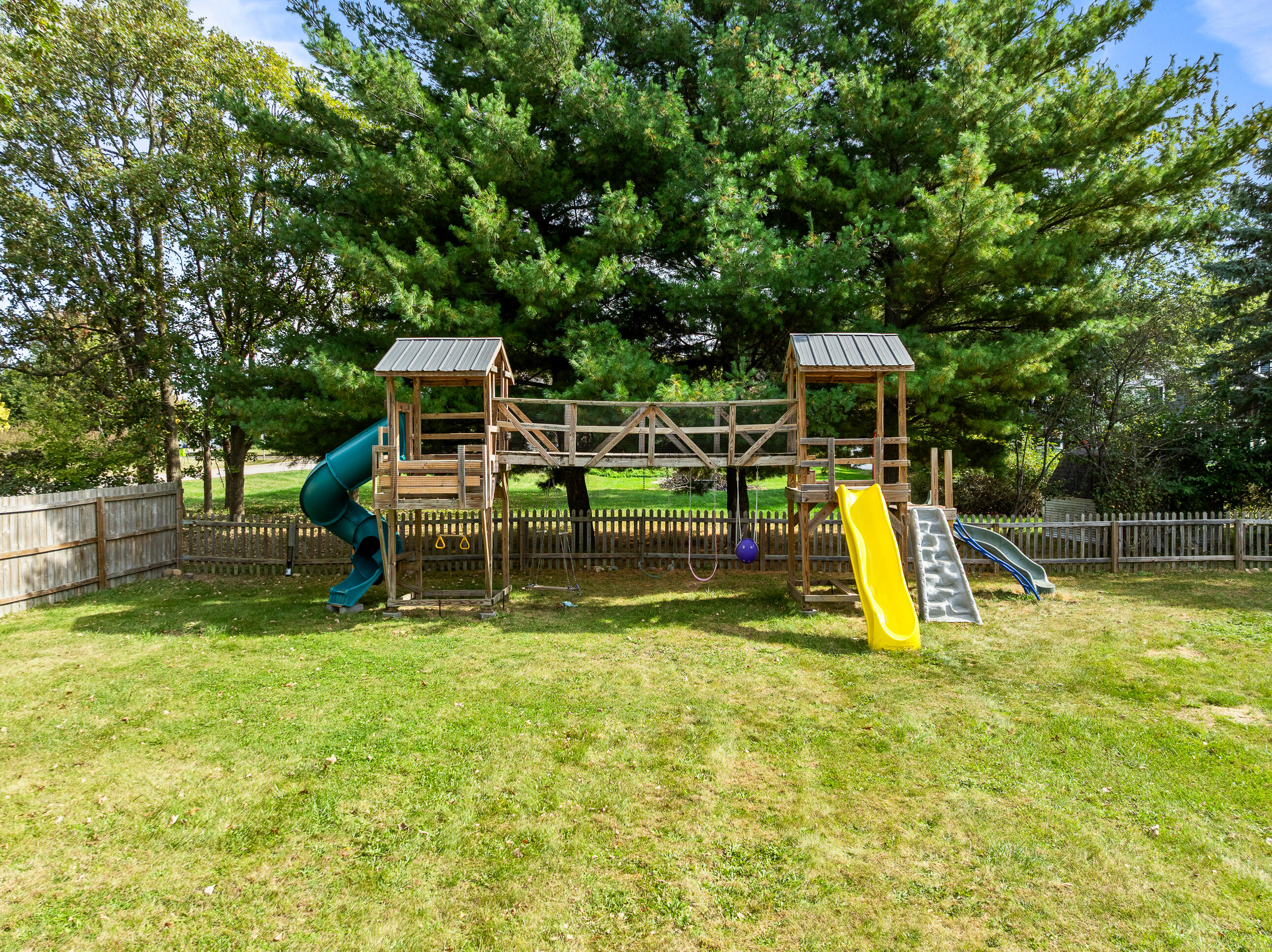 2212 Borg Road Delavan, WI 53115 - Photo 37 of 43 Large wooden play set with multiple slides and bridge walkway, all set within the fenced backyard with mature pine trees for a private backdrop.