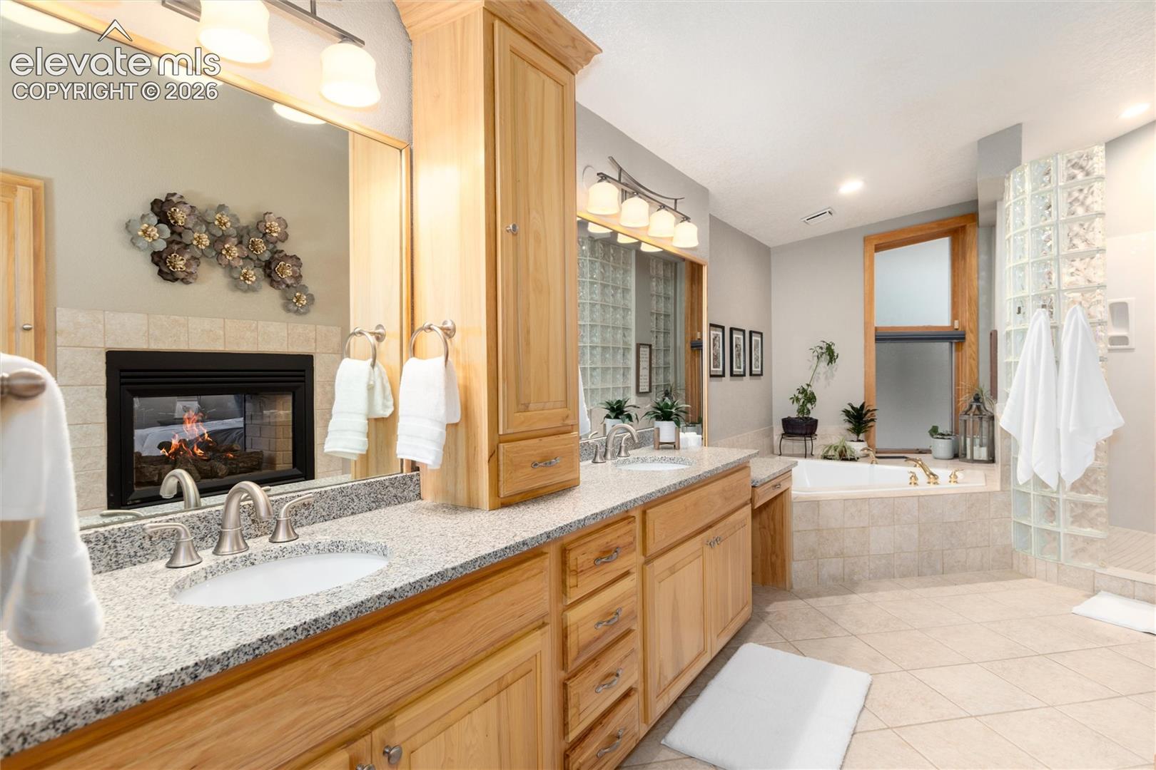 12730 Bridle Bit Road Colorado Springs, CO 80908 - Photo 15 of 48 Spacious bathroom featuring a dual vanity with granite countertops, built-in cabinetry, an oversized soaking tub, a glass block shower enclosure, and a fireplace