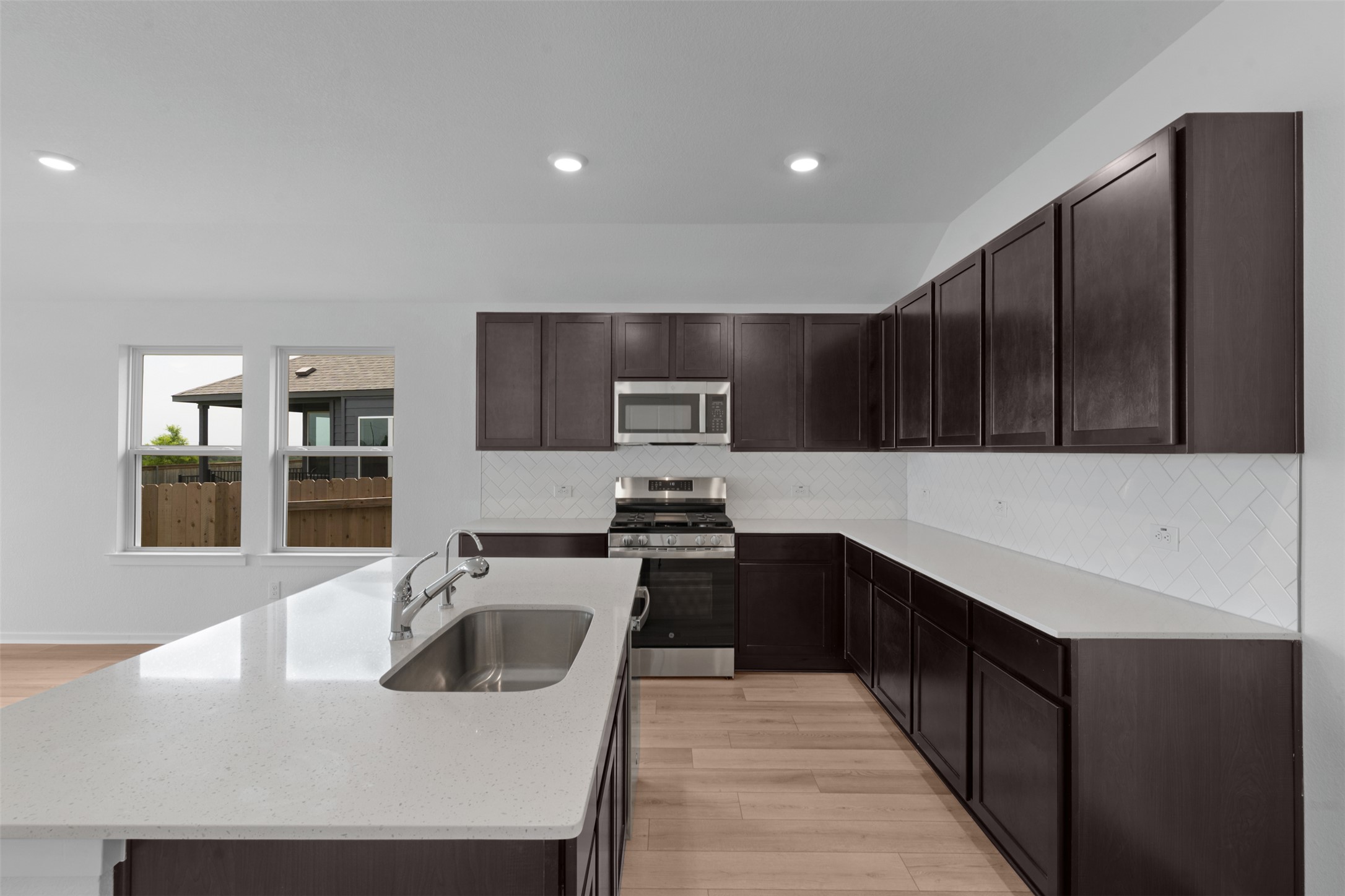 13701 Tucker Hedge Pass Elgin, TX 78621 - Photo 7 of 21 Kitchen featuring an island with an undermount sink, stainless steel appliances, dark wood-finish cabinetry, light-toned countertops, and a white subway tile backsplash