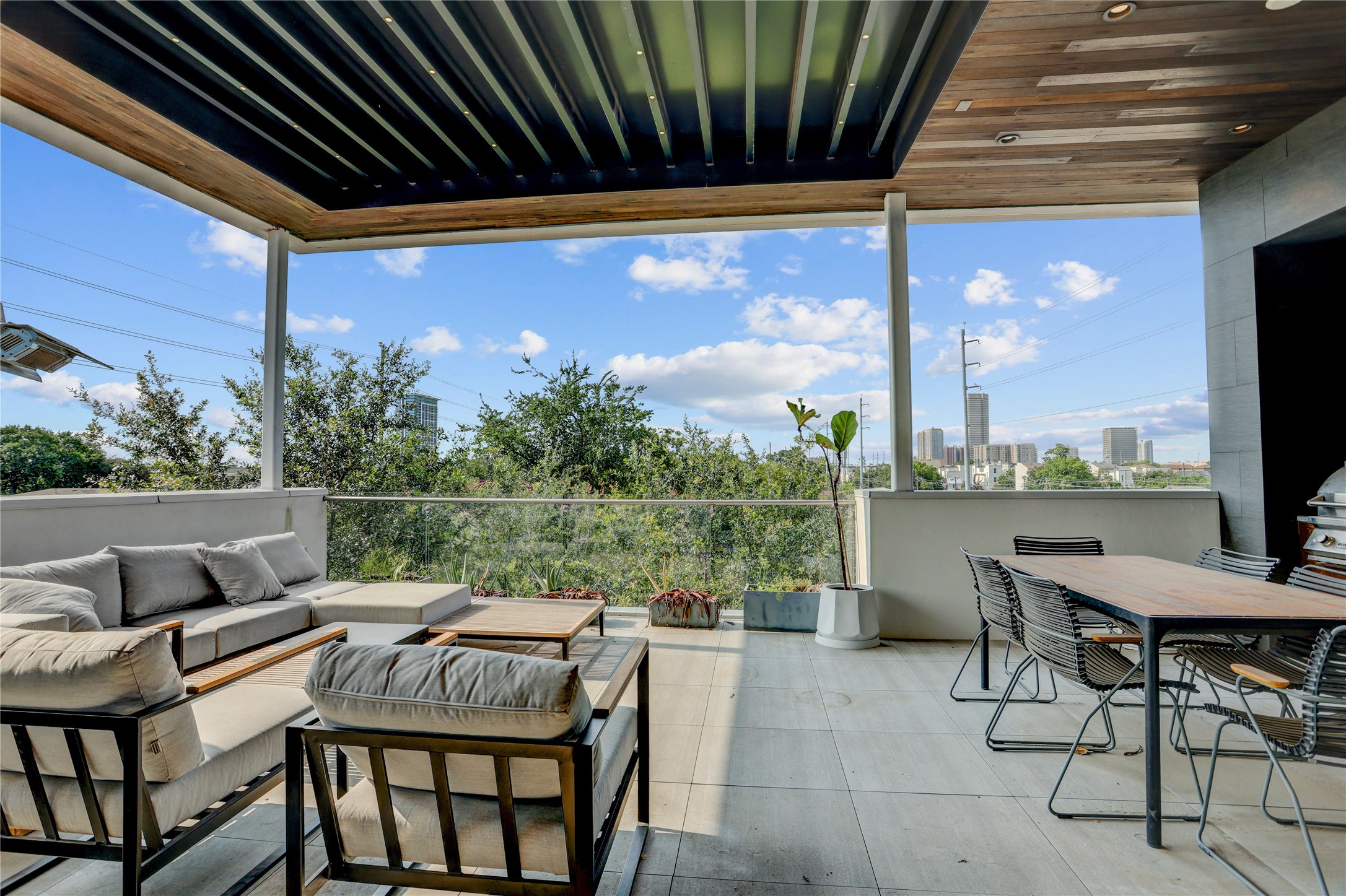 1807 Indiana Street Houston, TX 77006 - Photo 30 of 35 Rooftop Deck: Expansive fourth-floor covered terrace offering panoramic Houston skyline and treetop views, complete with a modern wood-paneled ceiling, glass-panel safety railings, an integrated outdoor kitchen setup, and ample space for both lounging and outdoor dining.