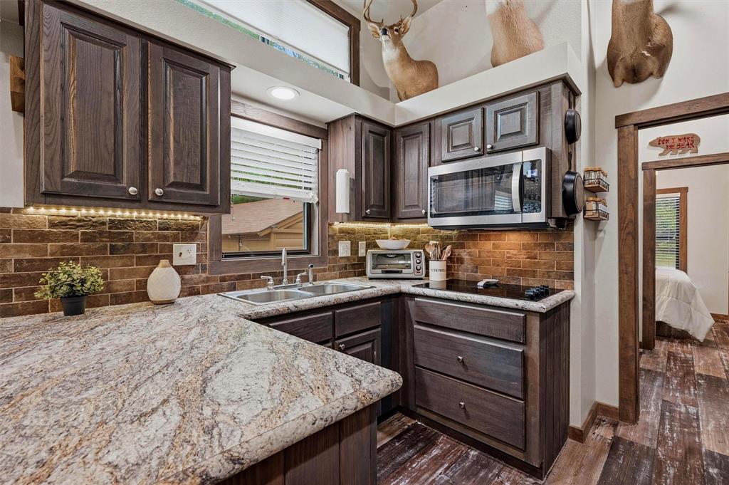 3958 Ranch Road Whitesboro, TX 76273 - Photo 13 of 40 Kitchen featuring an L-shaped counter with a double basin sink, dark wood-finish cabinetry, stainless steel microwave, integrated cooktop, and brick-style backsplash