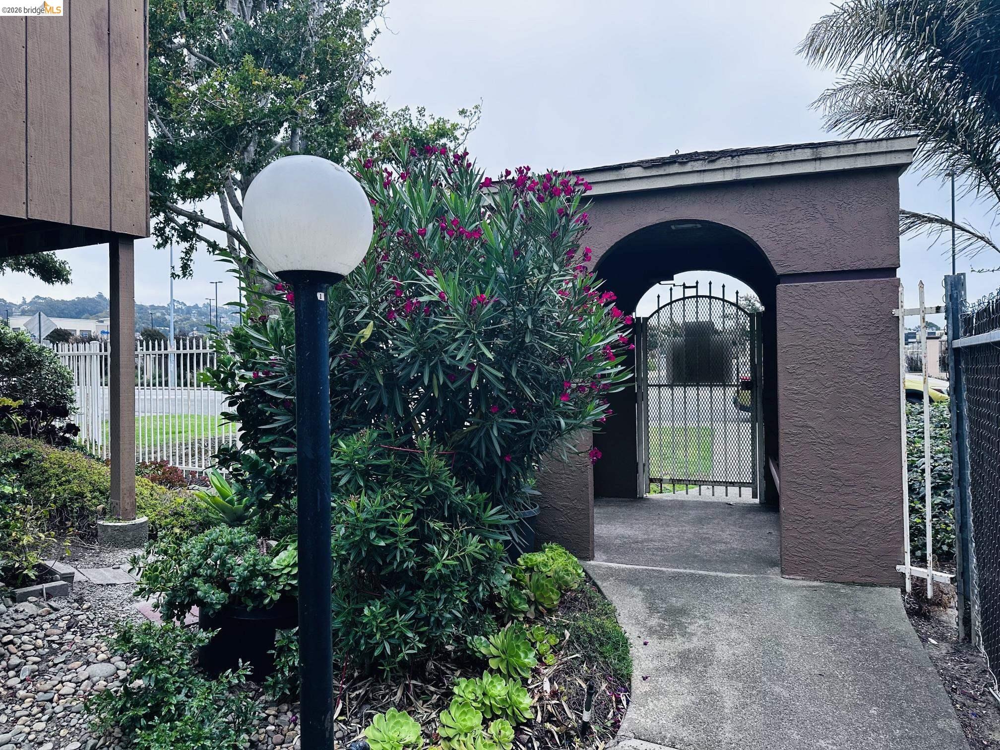 2121 Vale Road, Unit 17 San Pablo, CA 94806 - Photo 3 of 17 Arched entryway featuring an ornamental metal gate, a concrete walkway, mature flowering shrubs, and a path light