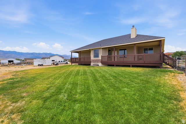 2567 West Rim Rock Drive Stockton, UT 84071 - Photo 50 of 64 Expansive green lawn with a tan exterior residence featuring a covered deck, dark brown railing, and an exterior chimney