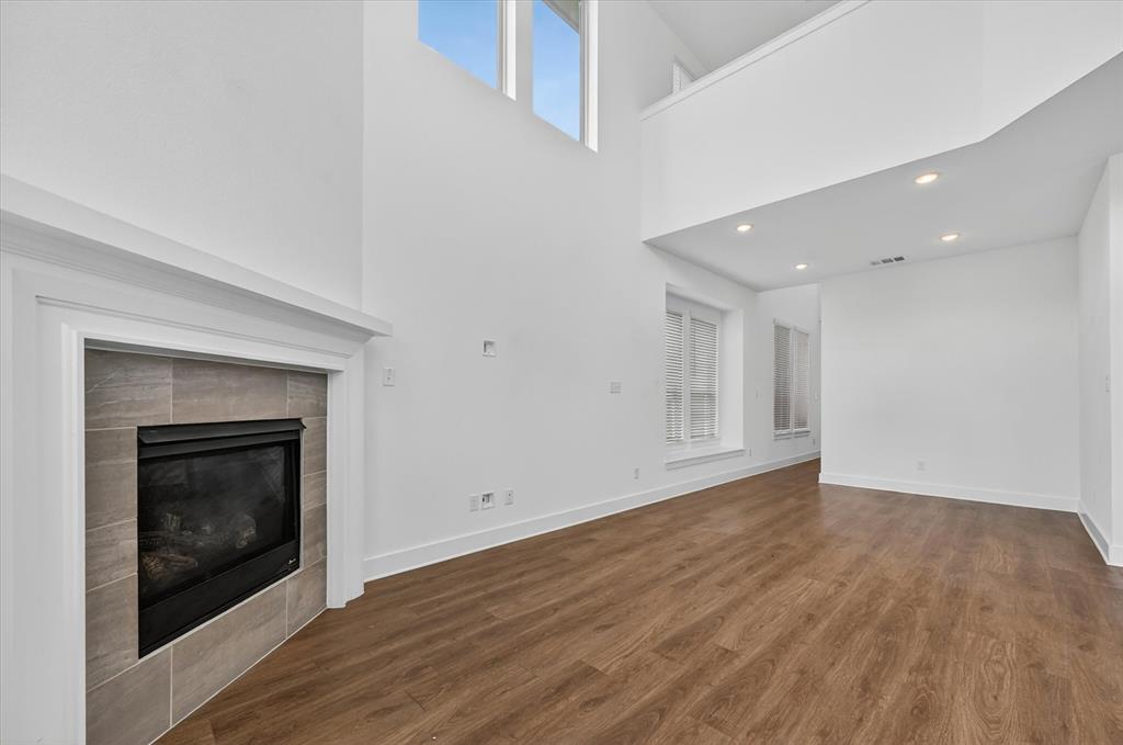 1153 Queensdown Way Forney, TX 75126 - Photo 7 of 26 Two-story living area featuring a fireplace with a tiled surround and white mantelpiece, wood-finish flooring, recessed lighting, and high windows providing natural light