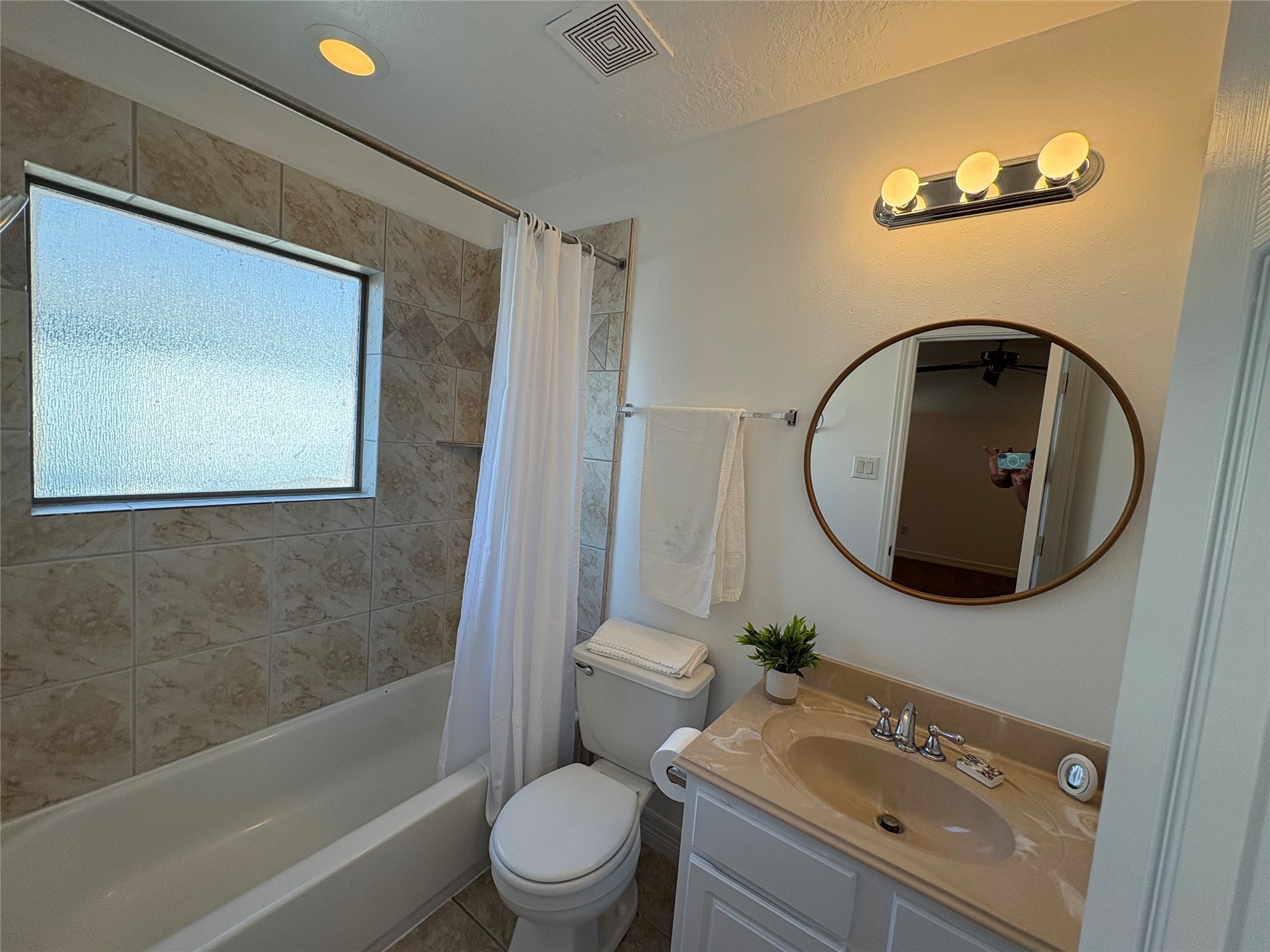 3710 Sherwood Lane, Unit 102 Houston, TX 77092 - Photo 14 of 27 Bathroom features a clean and simple design with a tiled shower and white curtain. It includes a round mirror above a vanity with a beige countertop, complemented by modern fixtures. There’s a small plant for a touch of greenery and a towel rack for convenience.