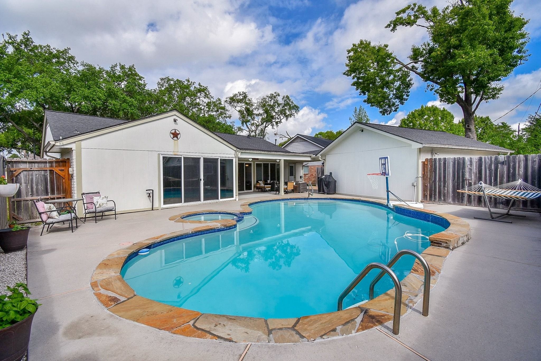 2930 Kevin Lane Houston, TX 77080 - Photo 13 of 50 This photo features a charming backyard with a spacious swimming pool, complete with a basketball hoop and adjacent hot tub. The area includes a cozy patio with seating for relaxation and entertainment. Lots of additional seating provide perfect spots for lounging. The setting is private with fencing and lush trees in the background.