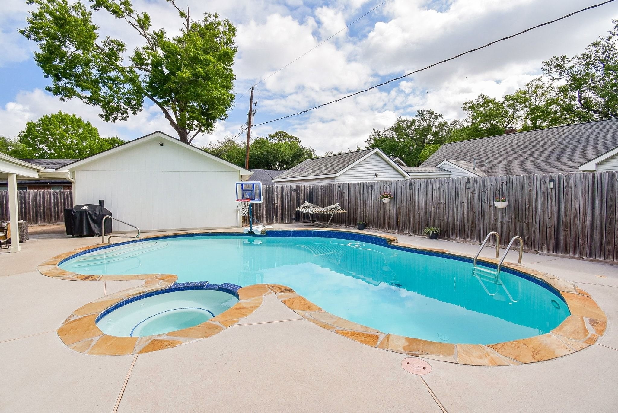 2930 Kevin Lane Houston, TX 77080 - Photo 14 of 50 This backyard features a spacious in-ground pool with a built-in spa, surrounded by a stone-trimmed patio. It offers privacy with a wooden fence and includes a basketball hoop and hammock for relaxation and recreation.