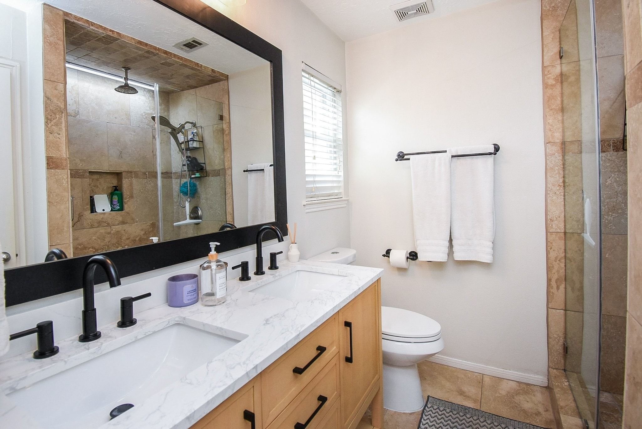 2930 Kevin Lane Houston, TX 77080 - Photo 48 of 50 This modern bathroom features a double vanity with a marble countertop, sleek black faucets, and a large mirror. The walk-in shower has elegant tile work and a glass door, while natural light filters through a window with blinds.