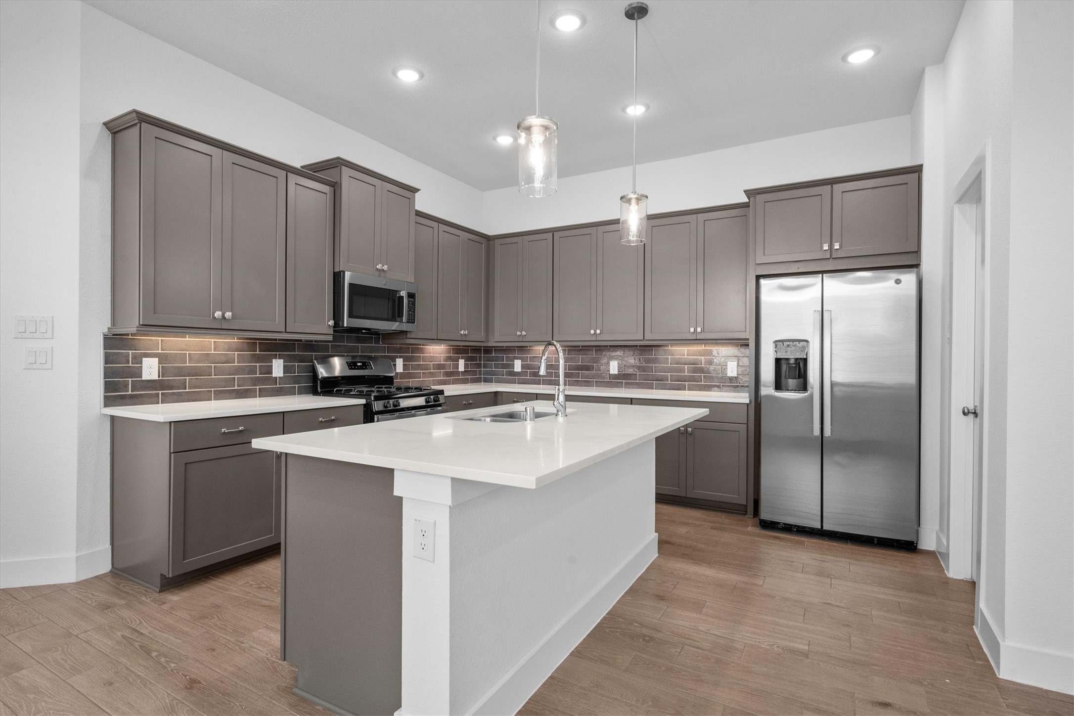120 Red Cascade Trail Willis, TX 77318 - Photo 11 of 37 This modern kitchen features sleek, dark cabinetry with a stylish subway tile backsplash and white countertops. It includes a central island with a sink, pendant lighting, and ample space for cooking and entertaining.