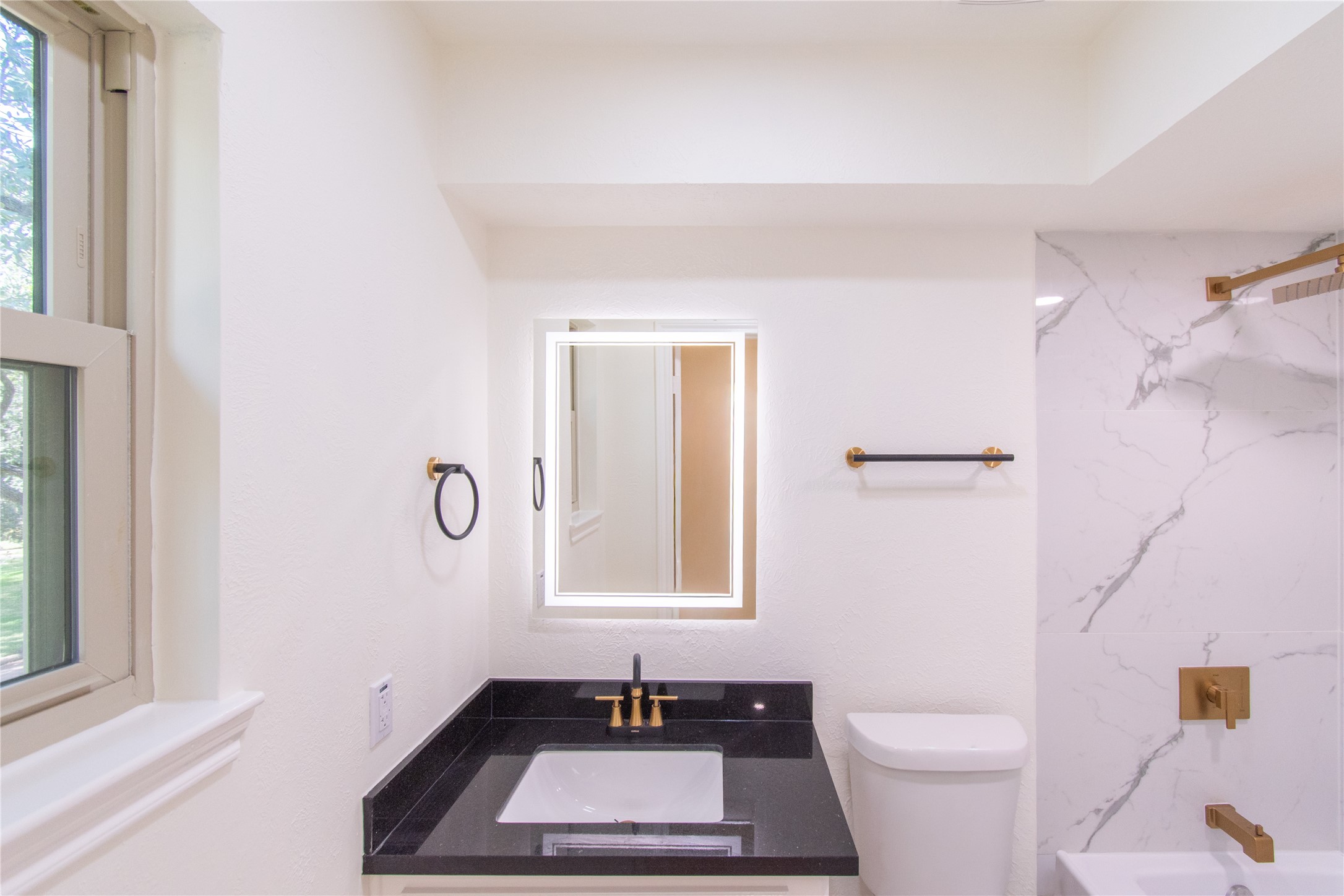727 Bunker Hill Road, Unit 43 Houston, TX 77024 - Photo 24 of 34 Modern and bright, featuring a sleek black countertop with a white sink and gold fixtures. It has a large, illuminated mirror and a stylish marble-patterned shower wall. Natural light streams in through a nearby window, enhancing the clean, contemporary look.